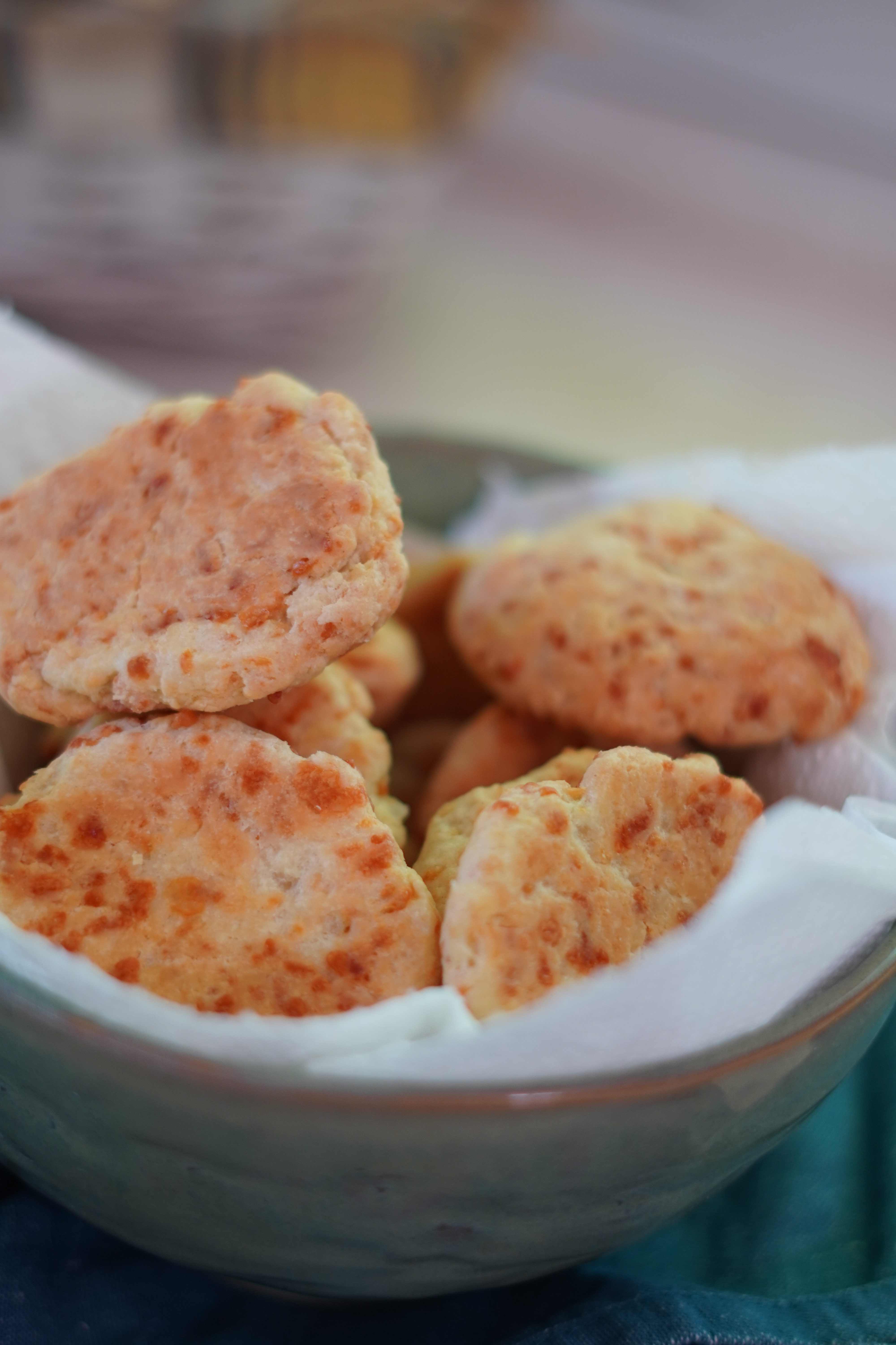 P&atilde;es de queijo pequenos e dourados, com pedacinhos de queijo vis&iacute;veis, empilhados em uma tigela de cer&acirc;mica clara forrada com um pano branco