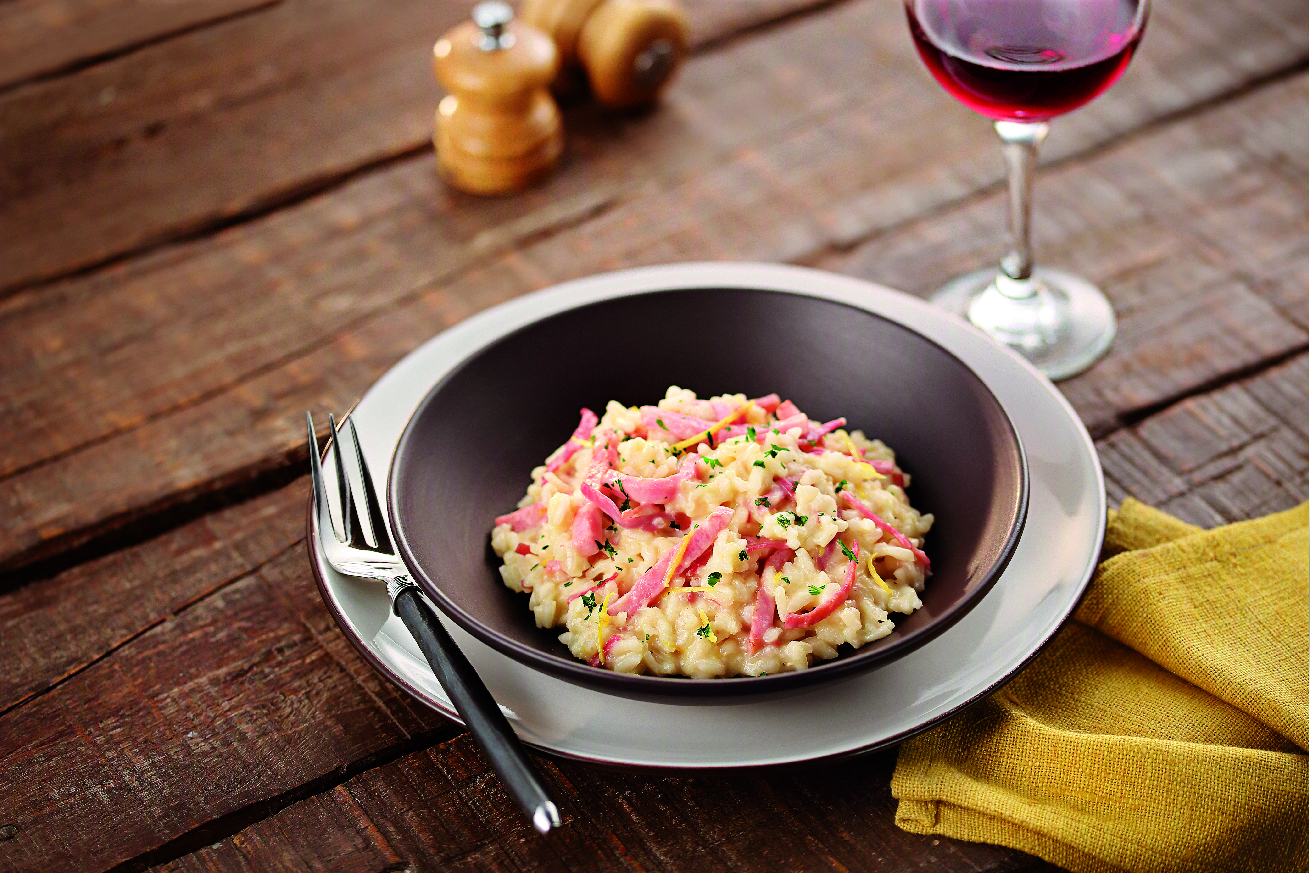 Risoto cremoso em tigela escura, com tiras de presunto rosa, raspas de lim&atilde;o amarelo e salsinha picada. Um garfo prateado, um guardanapo amarelo e uma ta&ccedil;a de vinho tinto completam a mesa de madeira r&uacute;stica