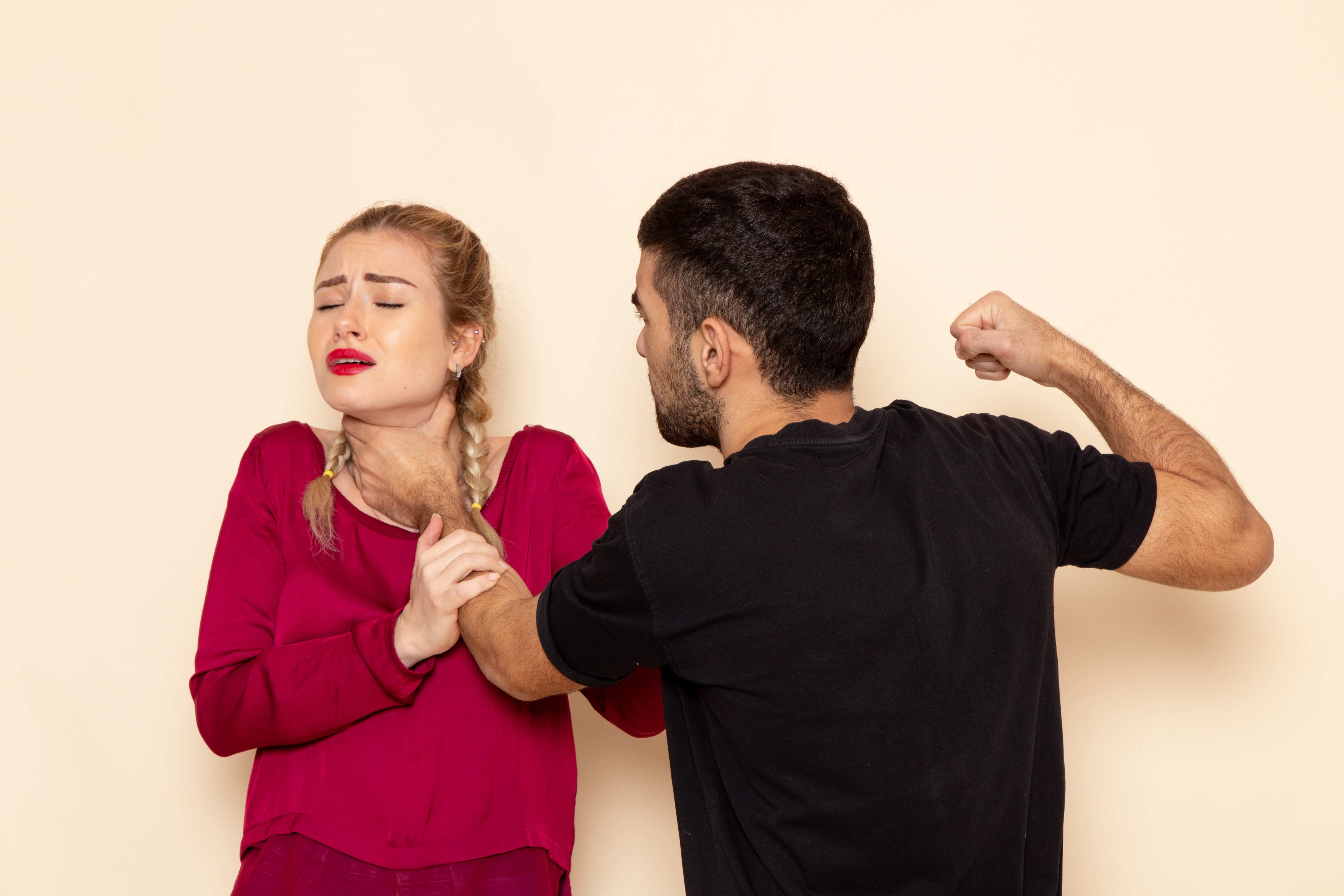 Homem de costas, com camiseta preta, amea&ccedil;a mulher loira de tran&ccedil;as, que veste blusa vinho e tem a garganta apertada por uma m&atilde;o, com express&atilde;o de dor e olhos fechados