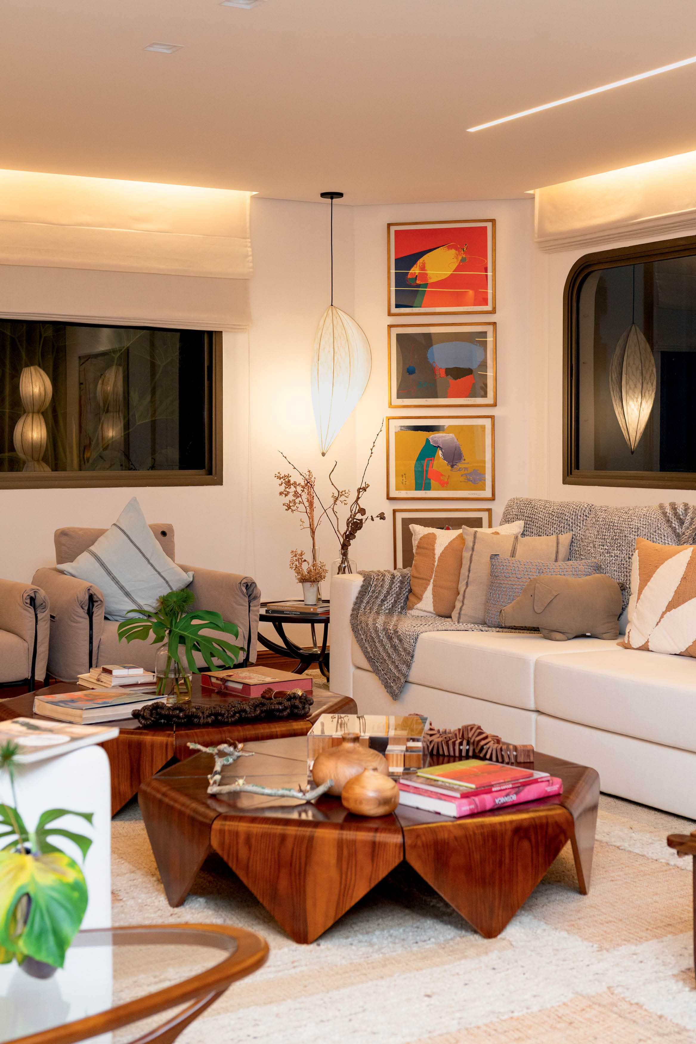 Sala de estar com sof&aacute; branco, poltronas bege, mesa de centro de madeira escura e tapete claro. Quadros abstratos coloridos na parede, lumin&aacute;rias pendentes e plantas decoram o ambiente aconchegante