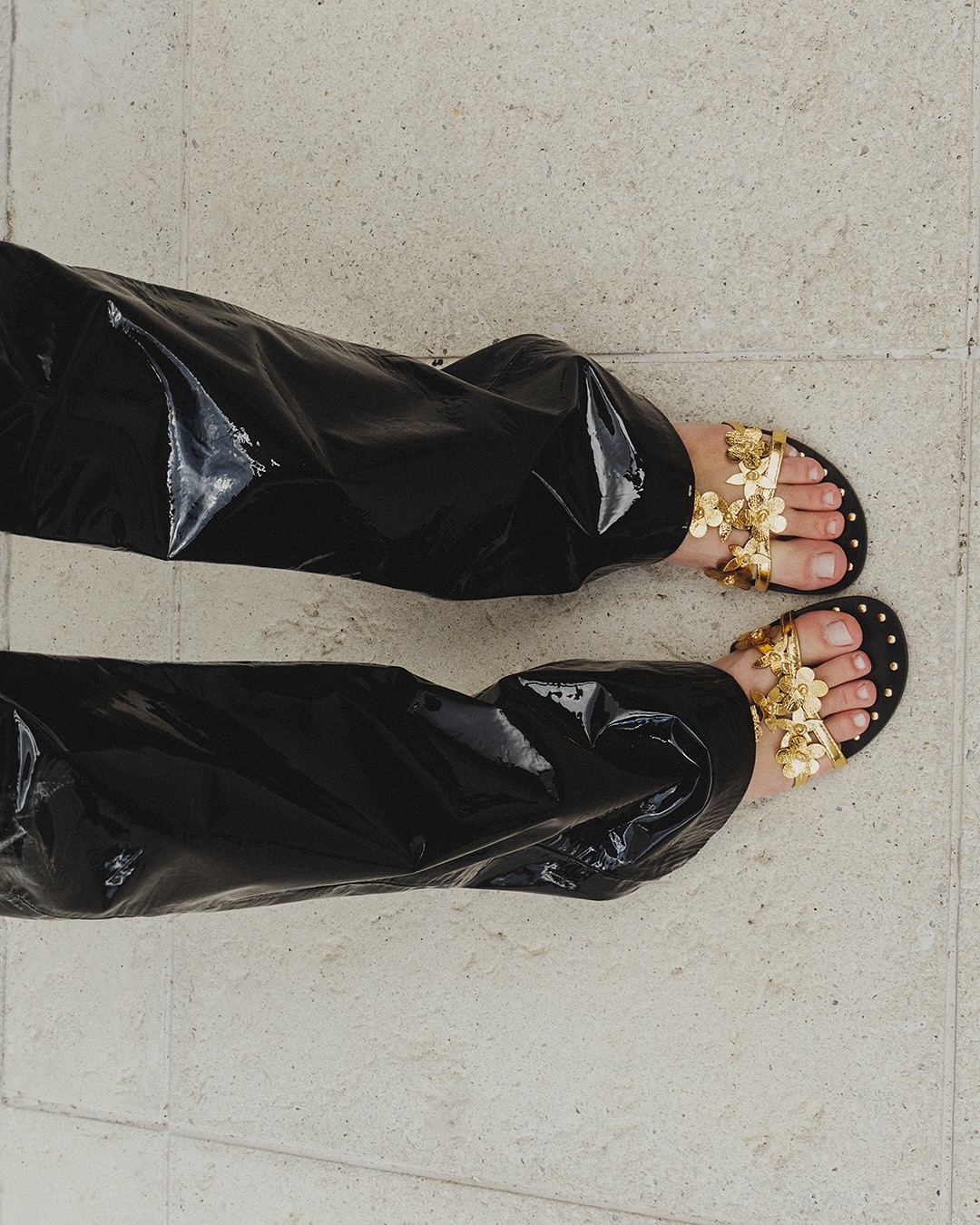 P&eacute;s de uma pessoa usando cal&ccedil;a preta de vinil e sand&aacute;lias rasteiras douradas com detalhes de flores, sobre um piso claro de cimento