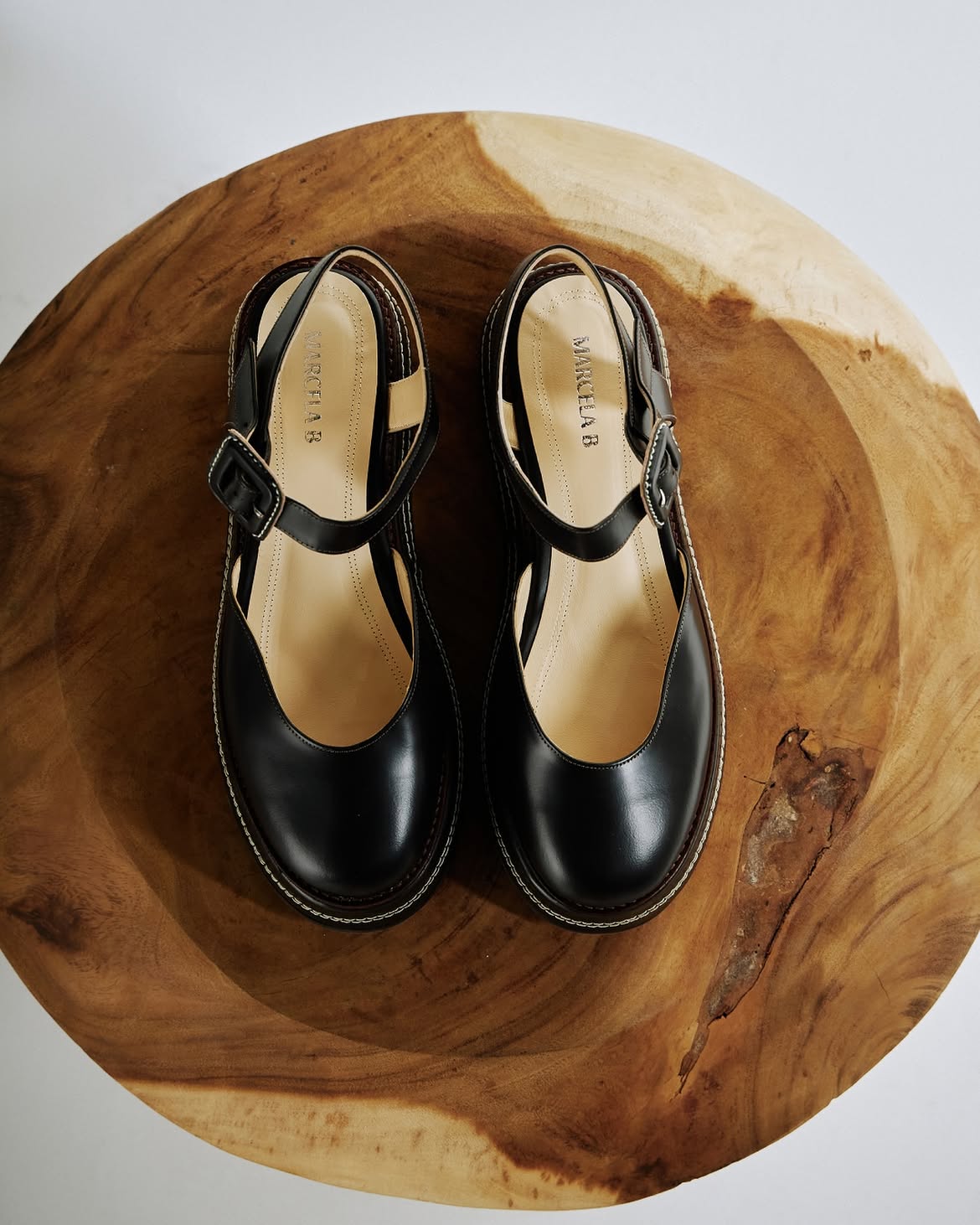 Par de sapatos pretos estilo Mary Jane com fivelas e solado grosso, sobre uma superf&iacute;cie de madeira r&uacute;stica com veios e n&oacute;s vis&iacute;veis