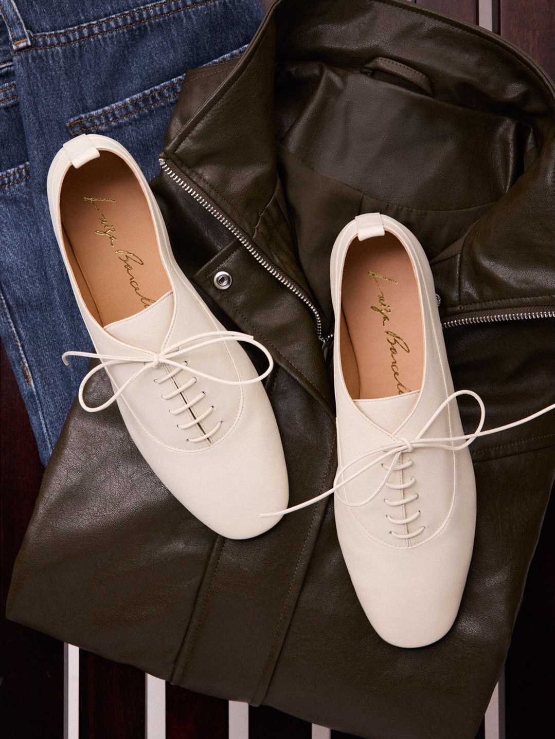 Dois sapatos oxford femininos brancos com cadar&ccedil;os finos sobre uma jaqueta de couro verde-militar e uma cal&ccedil;a jeans azul-escura, em um fundo escuro
