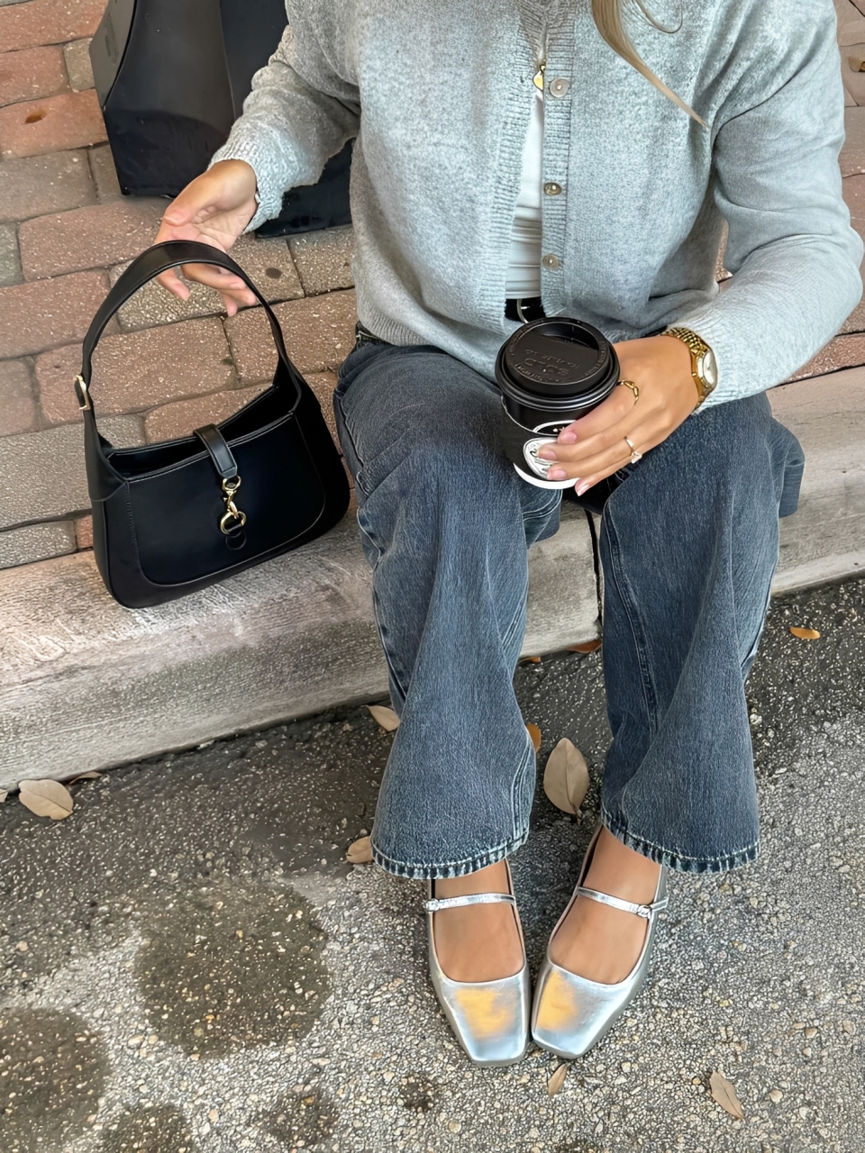 Pessoa sentada em um meio-fio, vestindo cardig&atilde; cinza, cal&ccedil;a jeans escura e sapatilhas prateadas. Segura uma bolsa preta e um copo de caf&eacute; descart&aacute;vel.