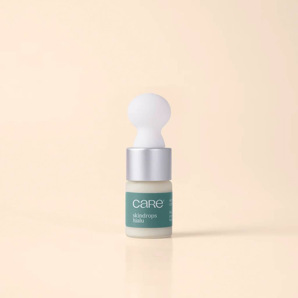 Frasco pequeno de vidro com conta-gotas, contendo um s&eacute;rum facial branco. O r&oacute;tulo verde-&aacute;gua exibe care e skindrops hialu em branco. A tampa prateada tem um bulbo branco esf&eacute;rico na ponta, sobre um fundo bege claro com reflexo do frasco