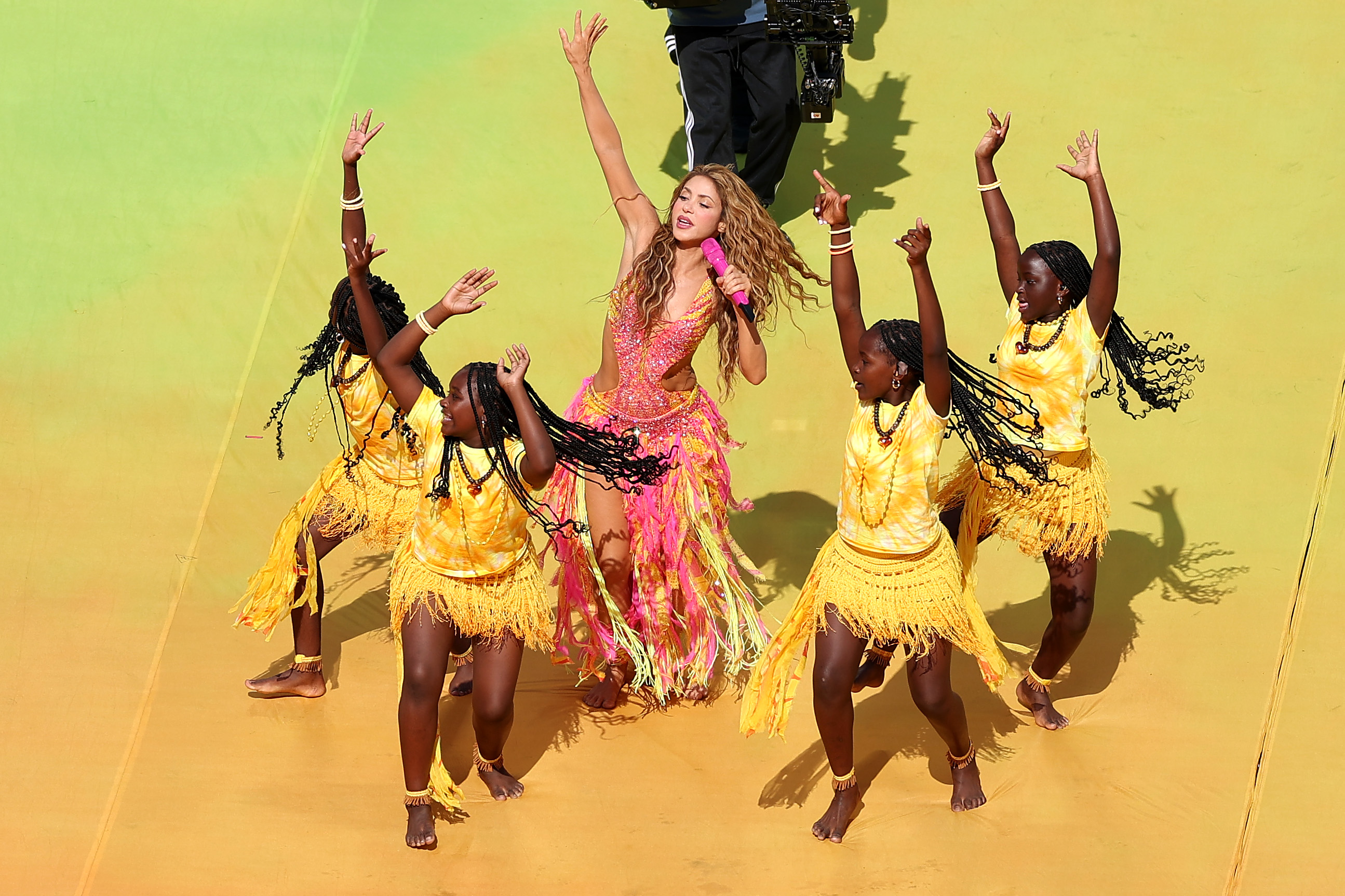 Shakira, de vestido rosa e laranja com franjas, canta em um microfone rosa, bra&ccedil;os erguidos, cercada por cinco dan&ccedil;arinas negras de tran&ccedil;as longas, vestindo blusas e saias de franjas amarelas, com os bra&ccedil;os levantados, em um palco amarelo e verde