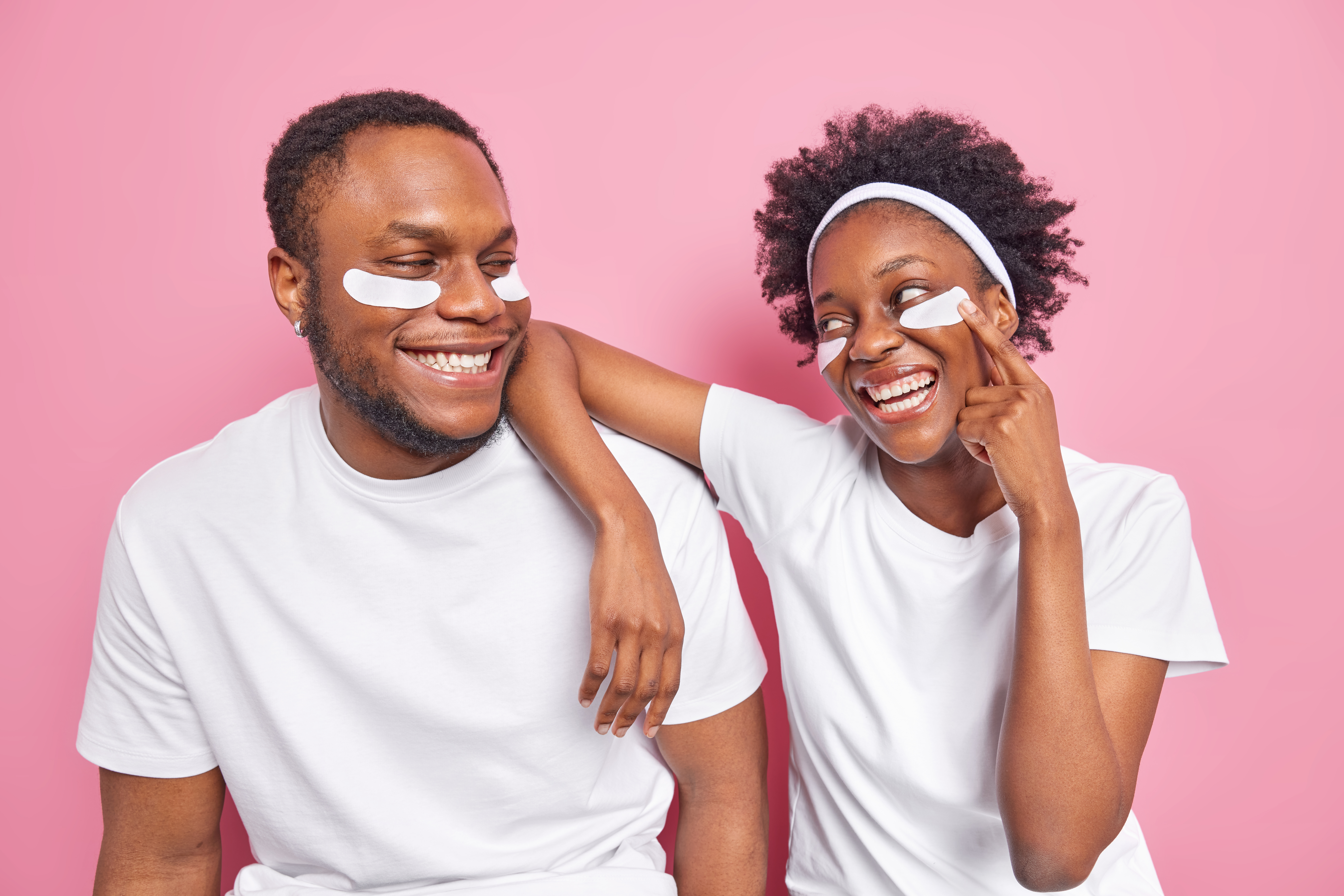 Um casal negro sorridente, ambos com camisetas brancas e m&aacute;scaras faciais brancas sob os olhos, em um fundo rosa. A mulher tem cabelo crespo e uma faixa branca, apontando para a m&aacute;scara do homem, que tem a barba aparada.