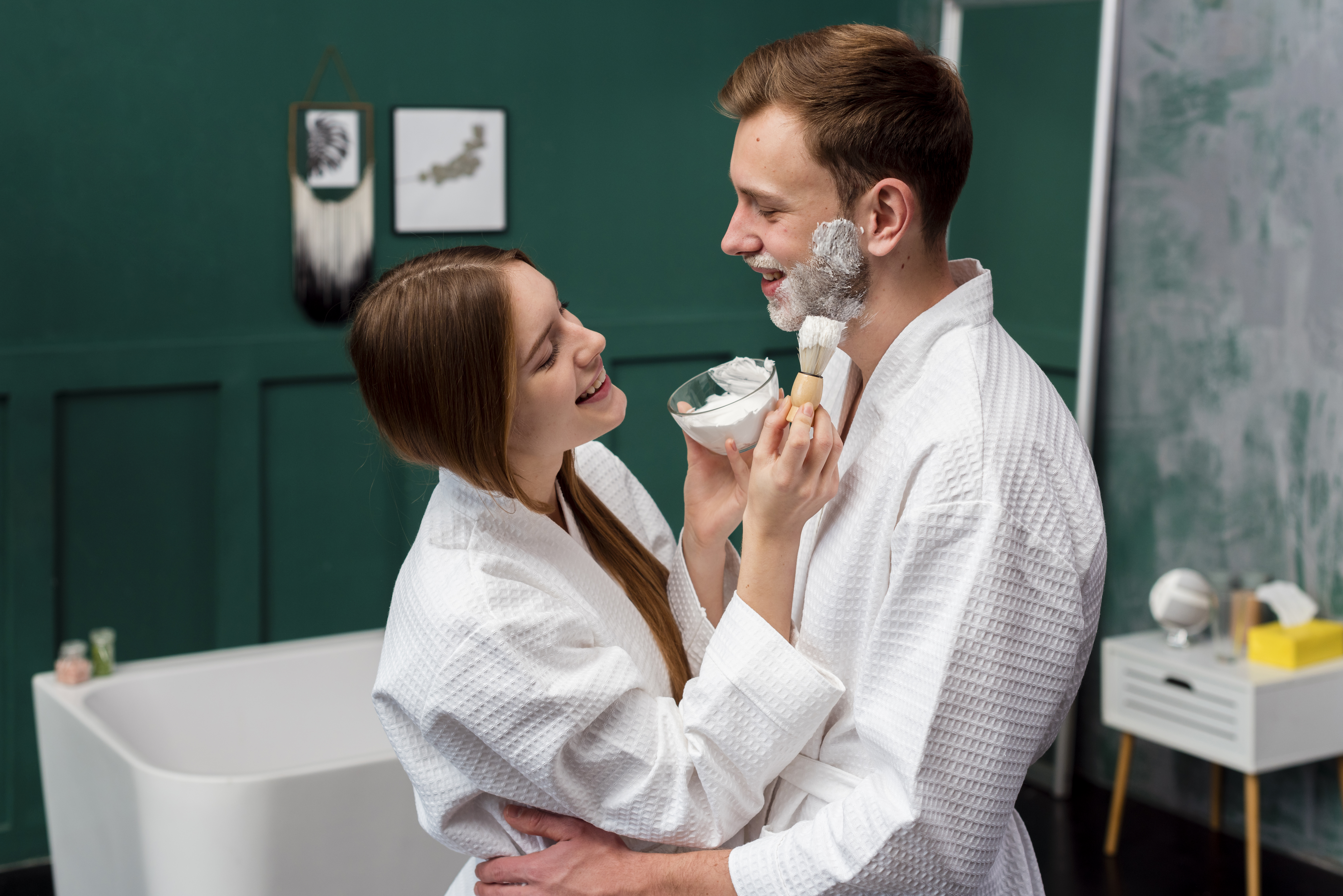Jovem casal sorridente em roupões brancos no banheiro. A mulher aplica espuma de barbear no rosto do homem com um pincel, segurando uma tigela. O homem tem espuma na bochecha e queixo, e a abraça pela cintura. Ao fundo, parede verde-escura, banheira branca e uma mesa de cabeceira.