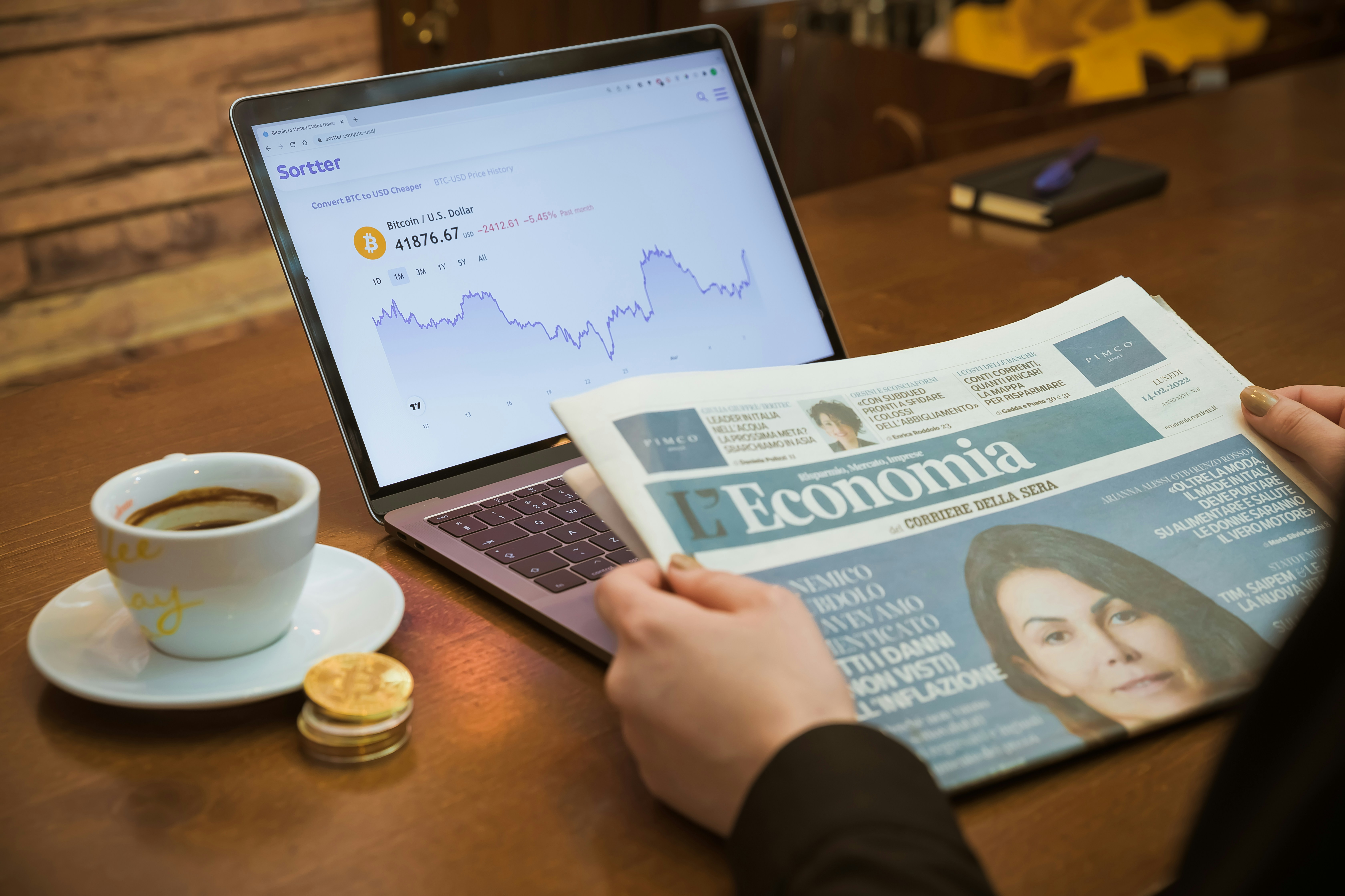 Pessoa lendo o jornal LEconomia com um laptop exibindo gráfico de Bitcoin e uma xícara de café ao lado, sobre uma mesa de madeira