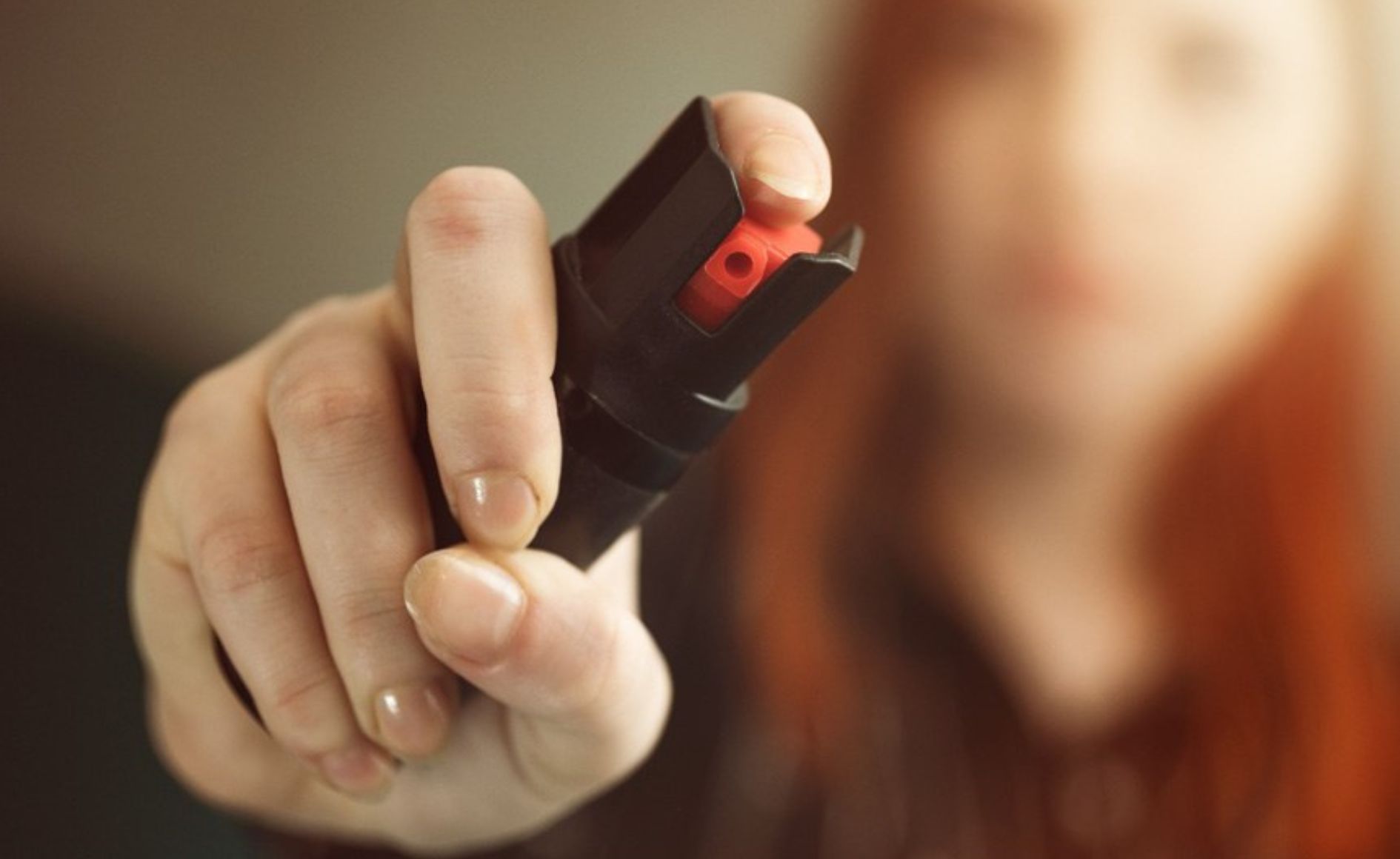 Mão feminina segurando um spray de pimenta preto com botão vermelho, apontando para frente, com o rosto da mulher desfocado ao fundo