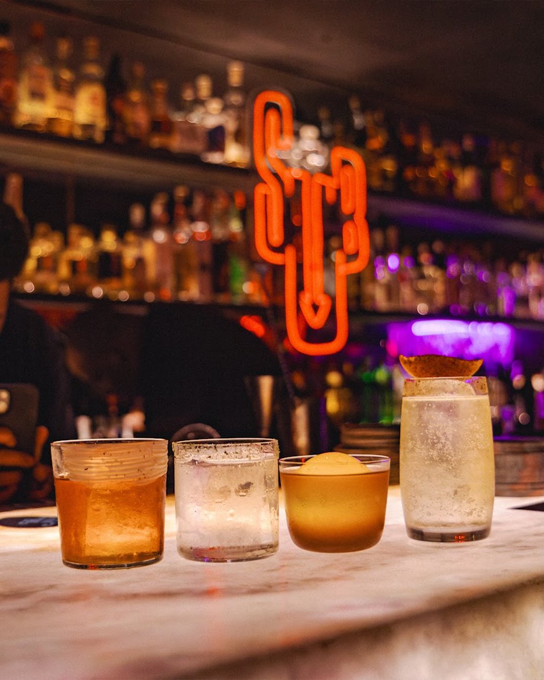 Quatro copos de bebidas variadas sobre um balc&atilde;o de m&aacute;rmore, com um letreiro neon laranja em formato de cacto e garrafas de bebidas em prateleiras iluminadas ao fundo