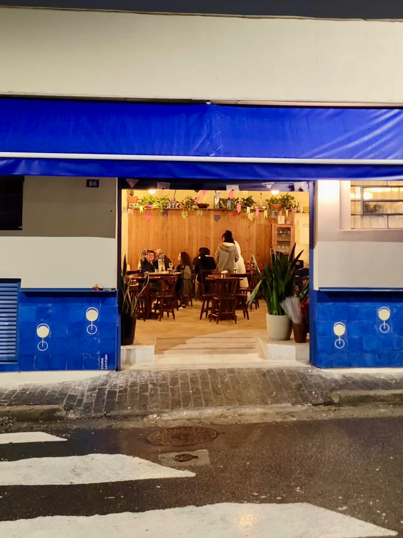 Fachada noturna de um restaurante com toldo azul e n&uacute;mero 86. Dentro, pessoas sentadas em mesas de madeira, com paredes de madeira e plantas decorativas. A cal&ccedil;ada tem piso de paralelep&iacute;pedos e a rua molhada reflete a luz.