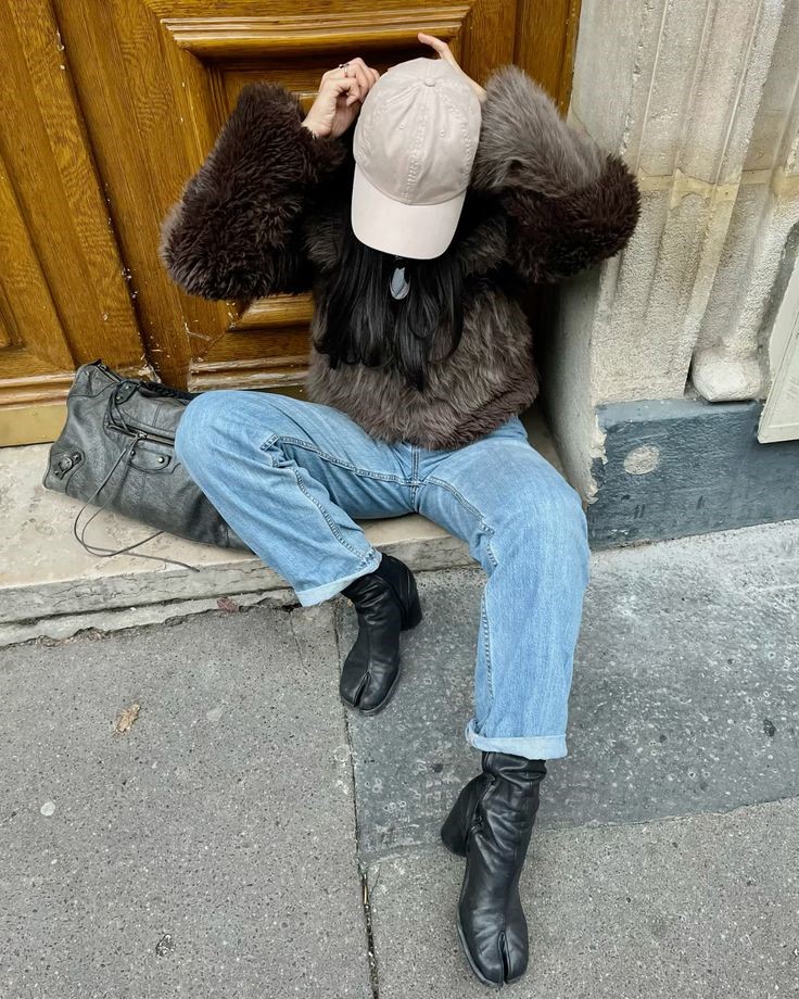 Pessoa sentada no ch&atilde;o, usando um casaco marrom de pelos, cal&ccedil;a jeans clara dobrada na barra, botas pretas com bico dividido e bon&eacute; bege. Uma bolsa preta est&aacute; ao lado, encostada em uma porta de madeira. O cen&aacute;rio &eacute; urbano, com cal&ccedil;ada de concreto e parede de pedra