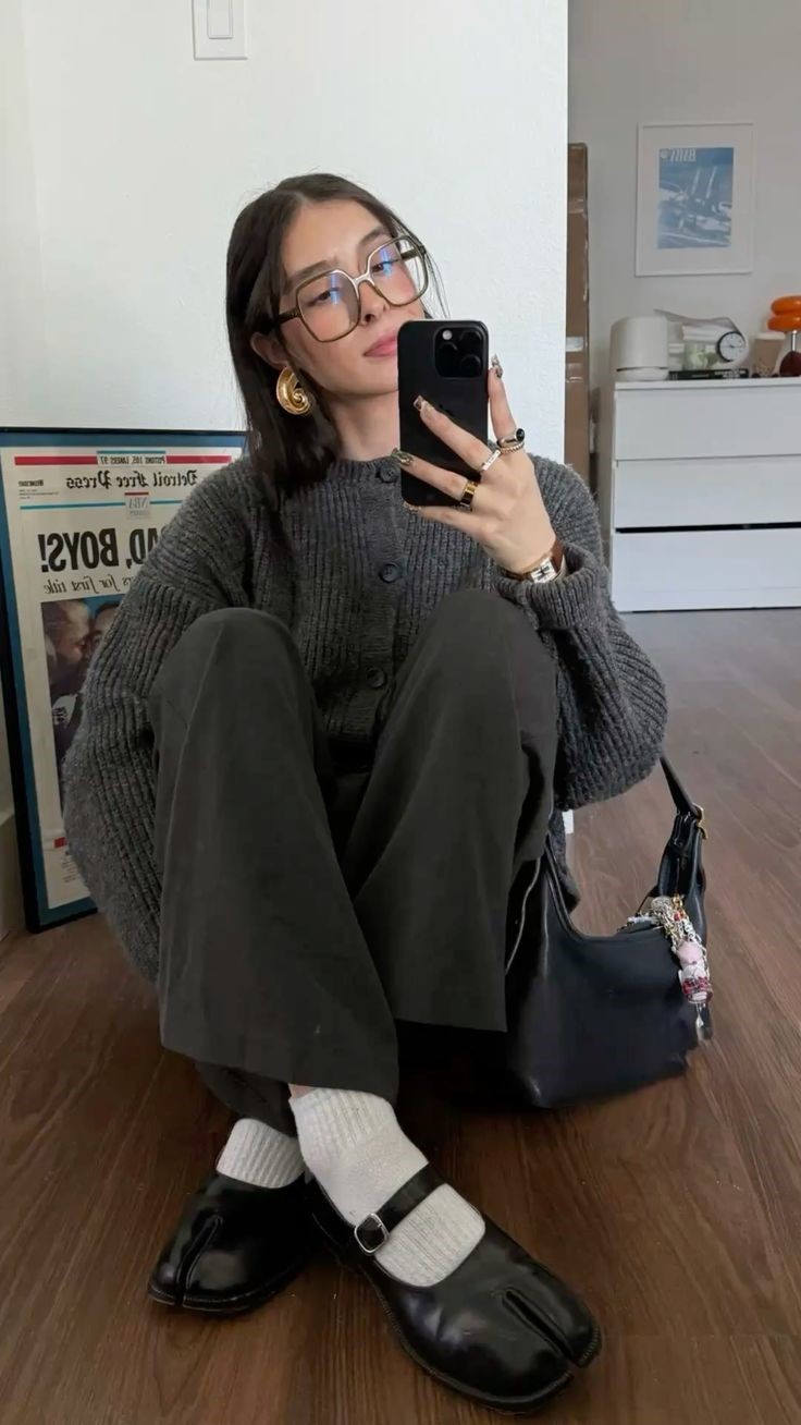 Mulher jovem com &oacute;culos grandes e brincos dourados, sentada no ch&atilde;o de madeira, tirando uma selfie. Ela veste um cardig&atilde; cinza, cal&ccedil;a escura, meias brancas e sapatos pretos com fivela. Ao fundo, um p&ocirc;ster e um m&oacute;vel branco com objetos decorativos