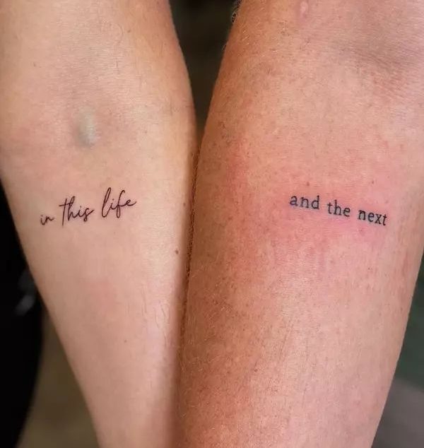 Dois antebra&ccedil;os lado a lado, um com a tatuagem in this life em caligrafia e outro com and the next em fonte serifada, simbolizando uni&atilde;o e continuidade