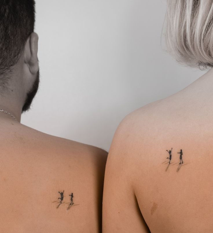 As costas de duas pessoas, um homem com barba e uma mulher de cabelo curto e loiro, exibindo tatuagens id&ecirc;nticas de duas figuras humanas em miniatura, uma com os bra&ccedil;os para cima e outra em p&eacute;, com sombras projetadas