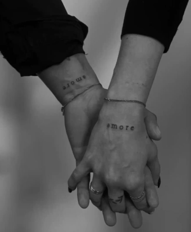 Duas pessoas de m&atilde;os dadas, em preto e branco. A m&atilde;o esquerda, com manga preta, tem a tatuagem amore no pulso, de cabe&ccedil;a para baixo. A m&atilde;o direita, com an&eacute;is e unhas escuras, tem a tatuagem amore no dorso. Ambas usam pulseiras finas.
