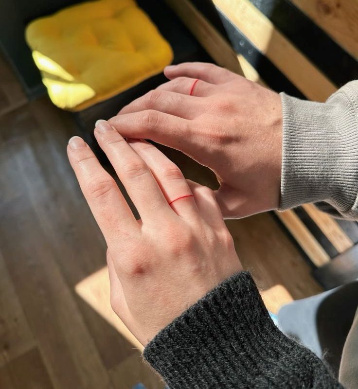 Duas m&atilde;os, uma feminina e uma masculina, exibem tatuagens de an&eacute;is vermelhos finos nos dedos anelares, simbolizando uni&atilde;o. A luz do sol incide sobre elas, criando sombras no fundo escuro com detalhes de madeira e um objeto amarelo