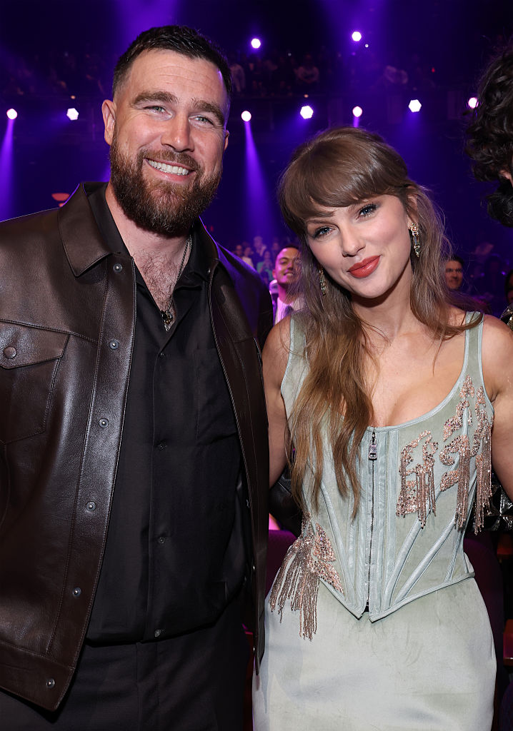O jogador Travis Kelce, barbudo e sorridente, veste jaqueta de couro marrom sobre camisa preta. Ao lado, a cantora Taylor Swift, com maquiagem marcante e batom vermelho, usa um top corset verde-&aacute;gua com detalhes brilhantes e franjas. Ambos est&atilde;o em um evento com ilumina&ccedil;&atilde;o roxa e p&uacute;blico ao fundo