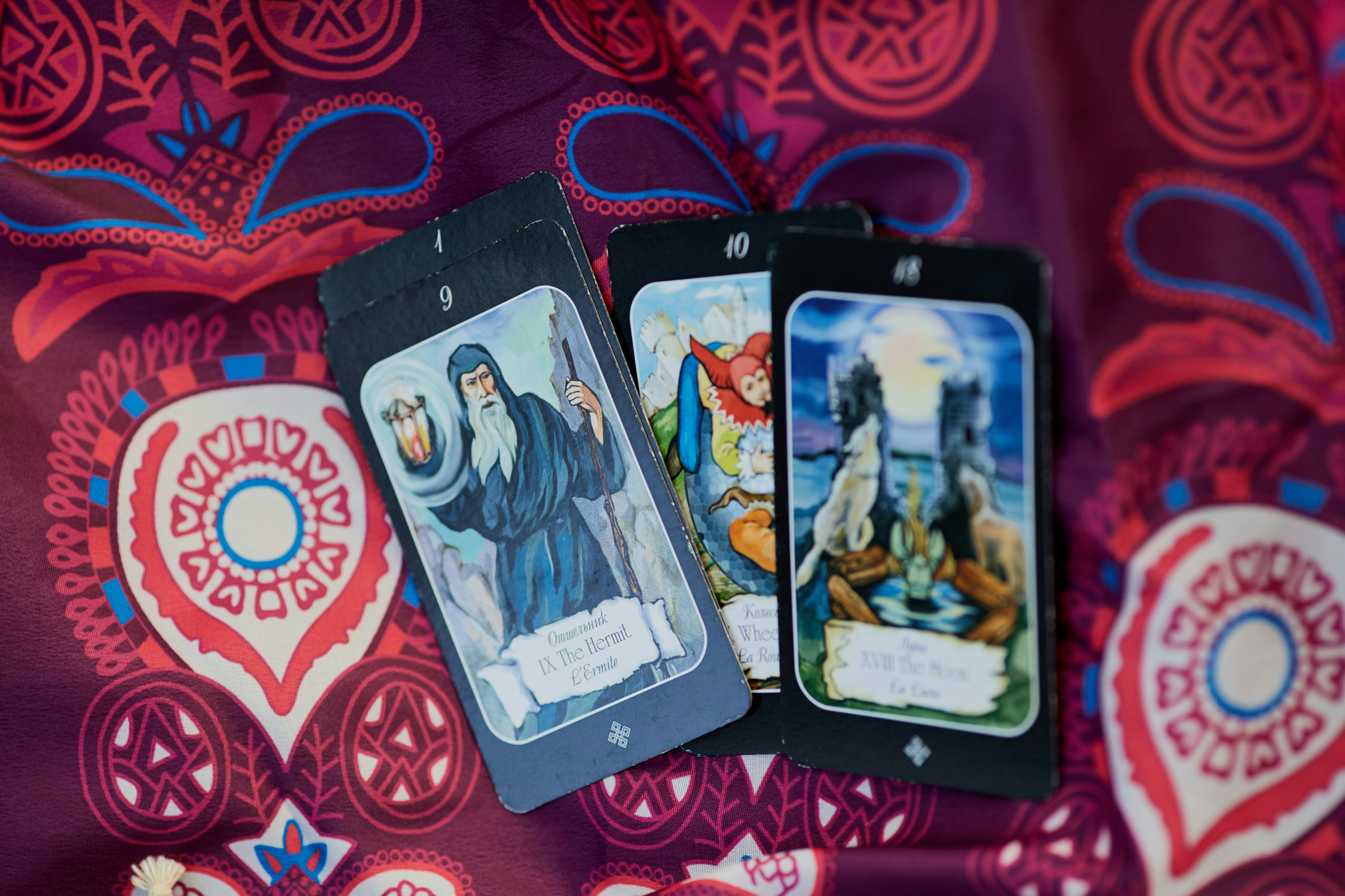 Três cartas de tarô sobre um tecido roxo com estampa paisley. A carta da frente mostra um eremita barbudo com cajado e lanterna, identificada como IX The Hermit.