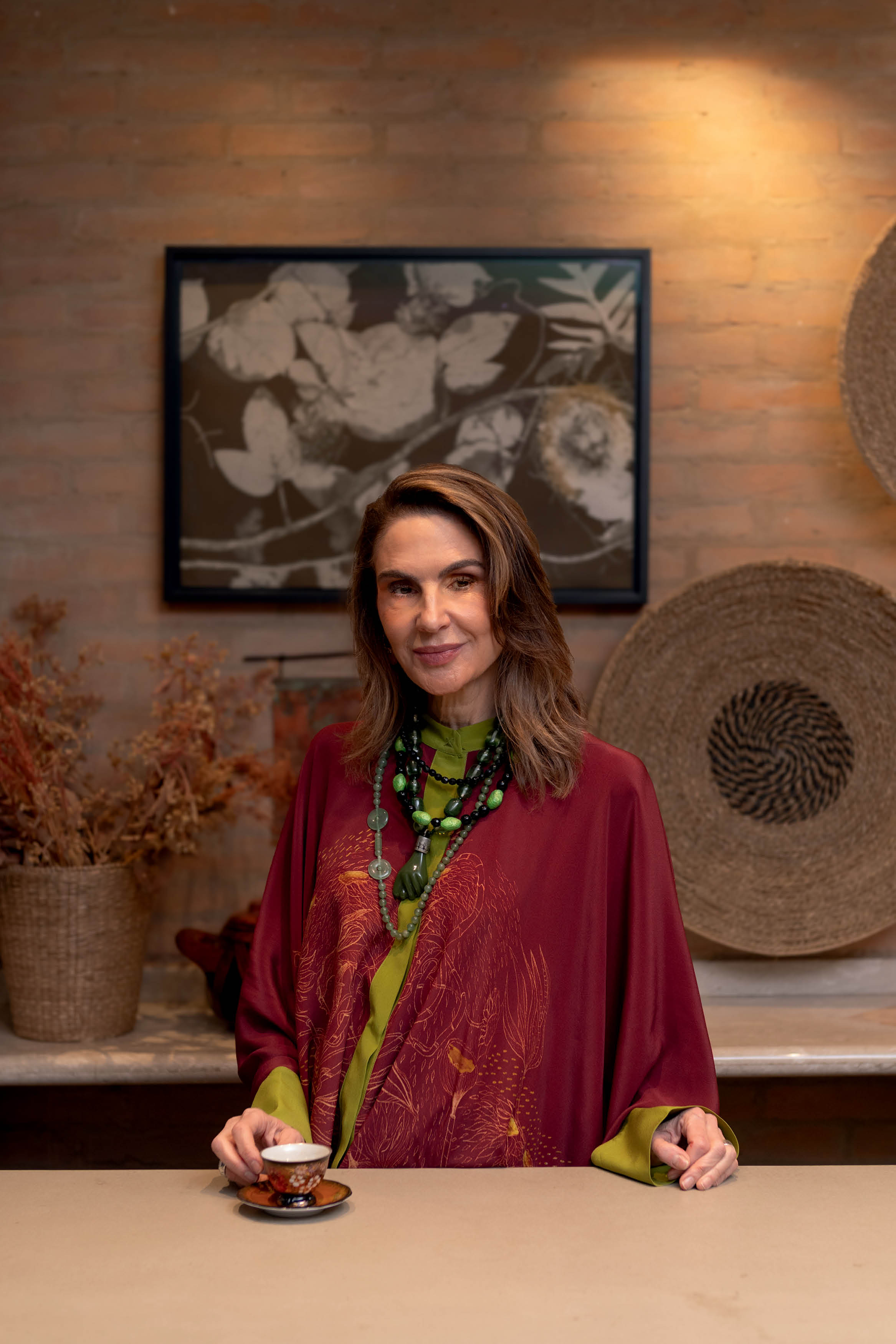 Mulher de meia-idade, cabelos castanhos e sorriso discreto, usando blusa verde-lim&atilde;o, kaftan vinho com estampa dourada e colares de contas verdes e pretas. Ela segura uma x&iacute;cara de ch&aacute; em um ambiente com parede de tijolos e quadros decorativos