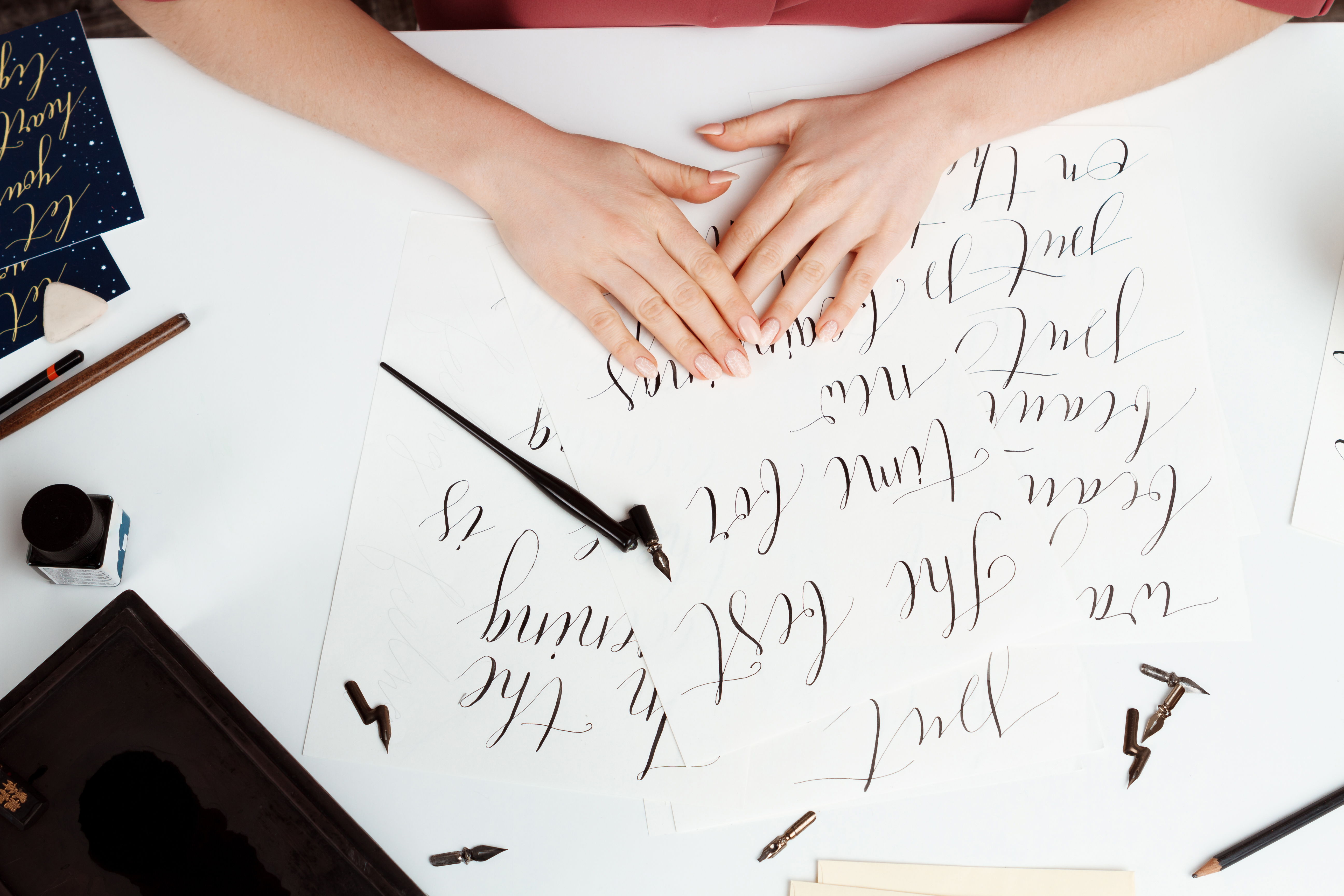 Mãos femininas com unhas claras sobre uma folha de papel branco com caligrafia em tinta preta. Ao redor, há um tinteiro, canetas de caligrafia, penas e um cartão azul com letras douradas.
