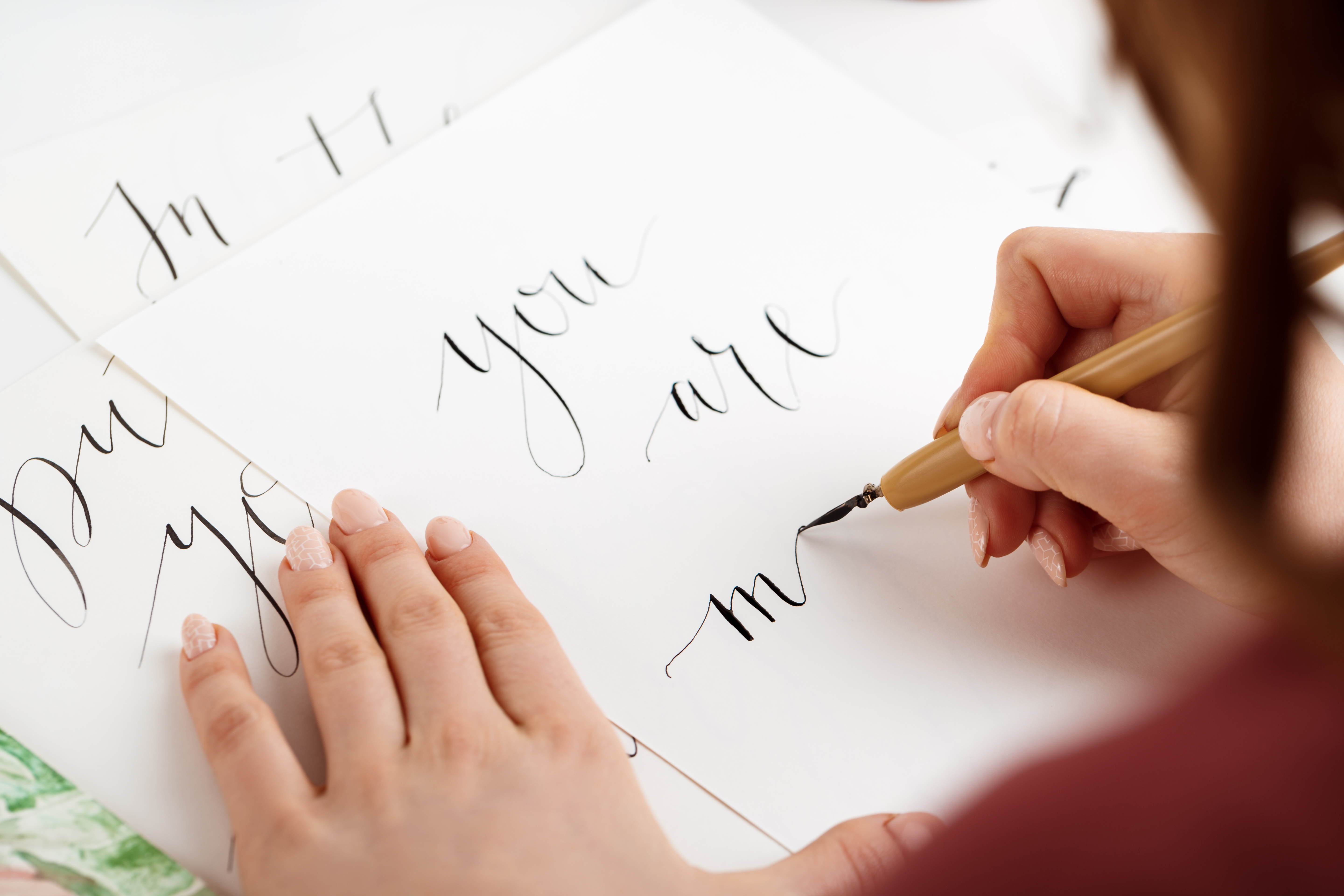 M&atilde;os femininas escrevendo caligrafia em papel branco com caneta-tinteiro. A m&atilde;o direita segura a caneta, formando a letra m, enquanto a esquerda apoia o papel. As unhas est&atilde;o pintadas de rosa claro, algumas com detalhes em renda. No papel, as palavras you are est&atilde;o escritas em caligrafia elegante