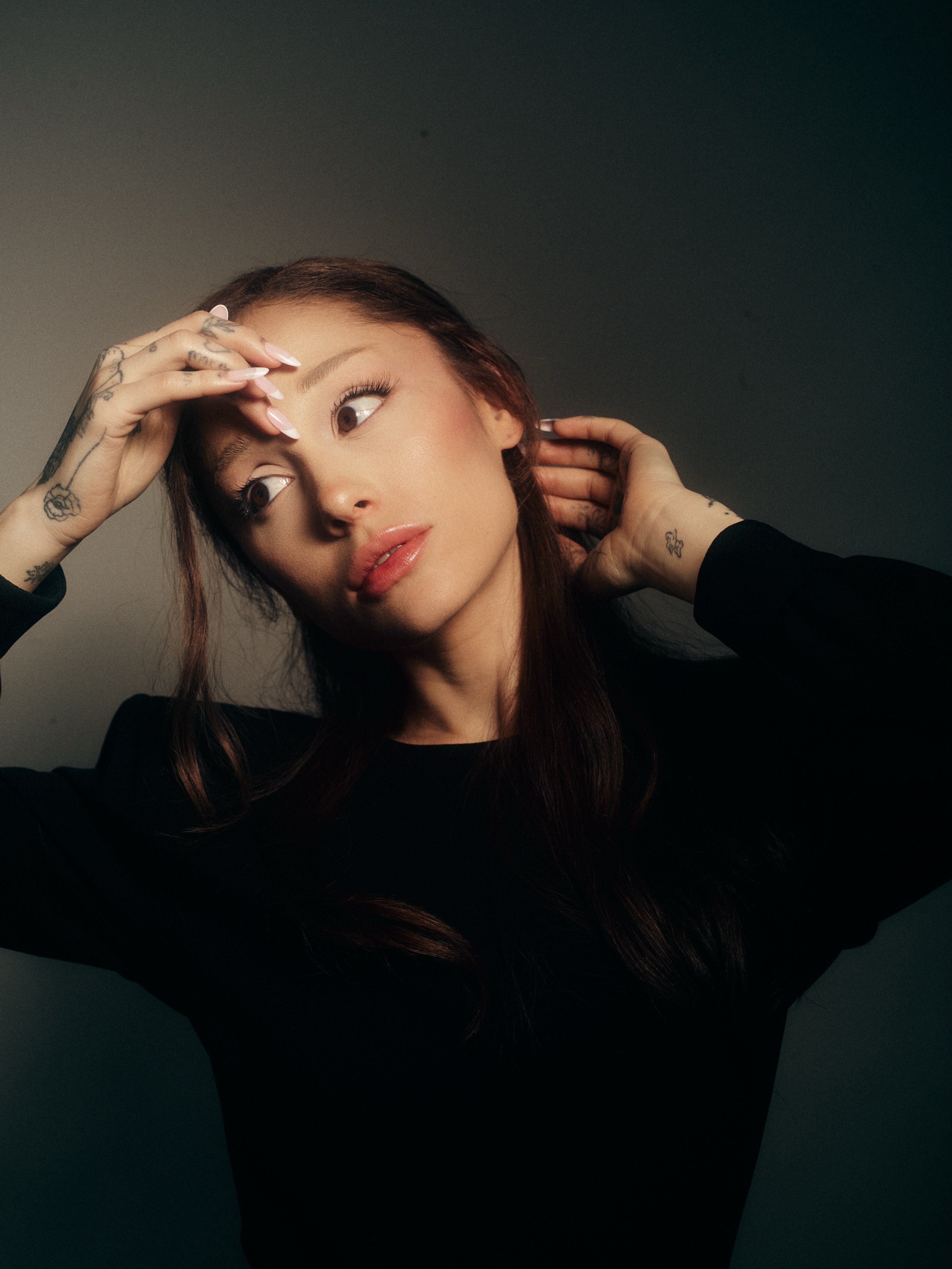 Cantora Ariana Grande com cabelo castanho solto, usando blusa preta, olhando para o lado esquerdo com express&atilde;o surpresa, m&atilde;os levantadas com unhas longas e tatuagens nos dedos e pulsos