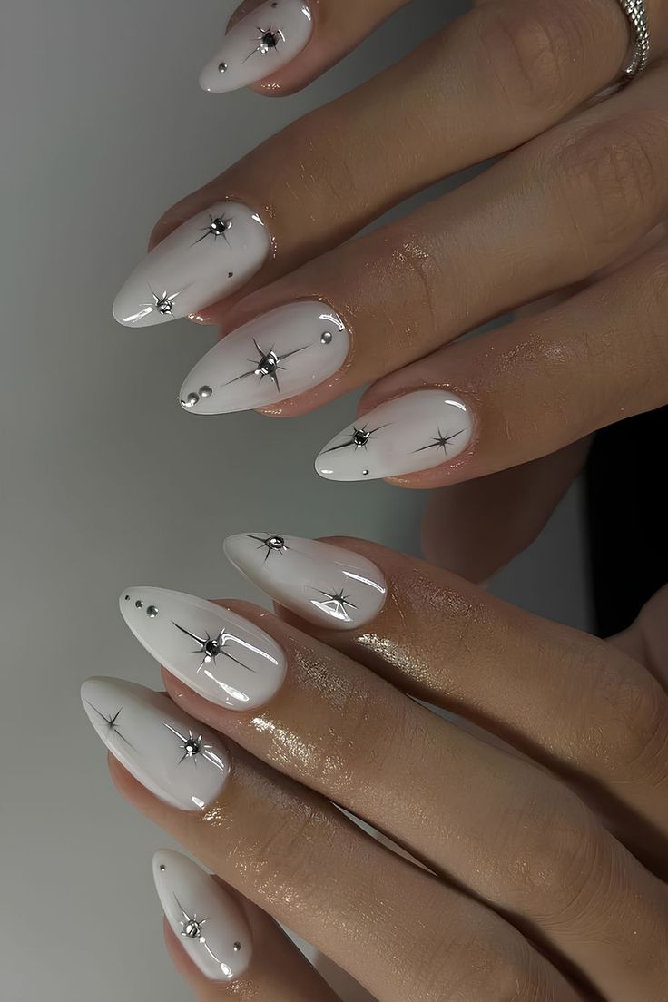 M&atilde;os com unhas amendoadas longas e brancas, decoradas com pedras prateadas e desenhos de estrelas pretas, em fundo cinza claro