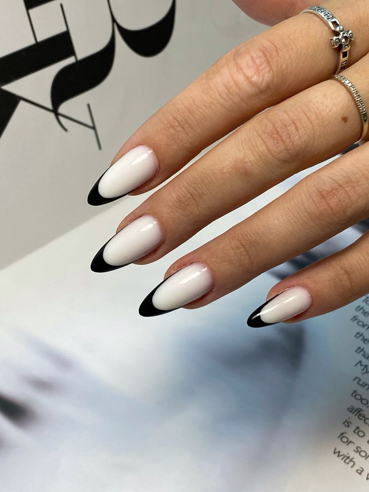 M&atilde;o feminina com unhas amendoadas em esmalte branco leitoso e pontas pretas, estilo francesinha invertida. Dois an&eacute;is prateados adornam o dedo anelar, um com detalhes em cruz e outro liso. A m&atilde;o est&aacute; sobre uma superf&iacute;cie branca com texto impresso em preto ao fundo