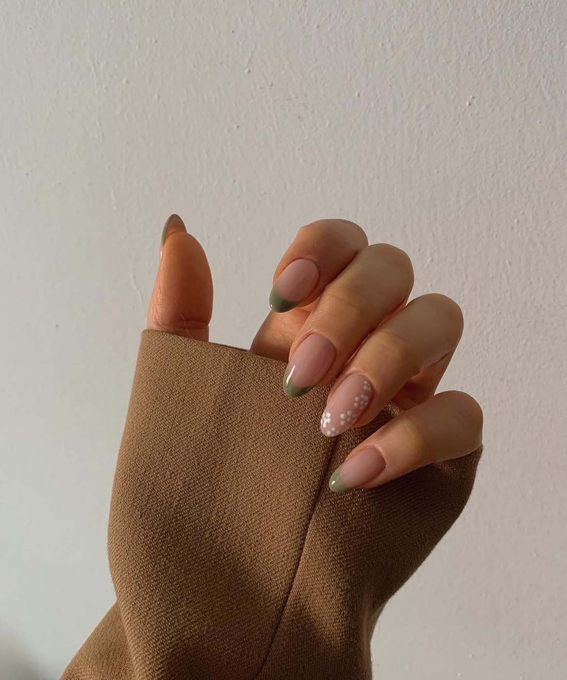M&atilde;o feminina com unhas ovais, esmalte nude e pontas verde-oliva. O dedo anelar tem pequenas flores brancas, e a m&atilde;o emerge de uma manga marrom de tecido texturizado, contra uma parede branca