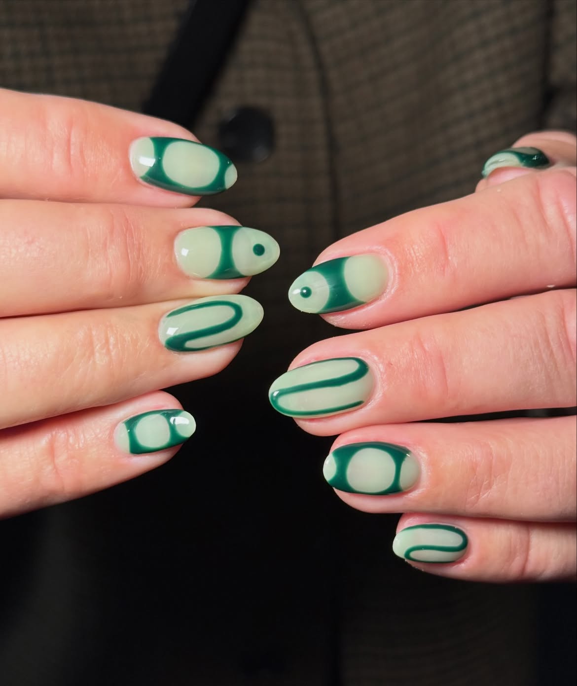 M&atilde;os femininas com unhas ovais decoradas em verde-escuro e verde-claro. Os desenhos variam entre c&iacute;rculos, semic&iacute;rculos e linhas onduladas, com um ponto verde-escuro no centro de algumas unhas. O fundo &eacute; um tecido marrom quadriculado e escuro