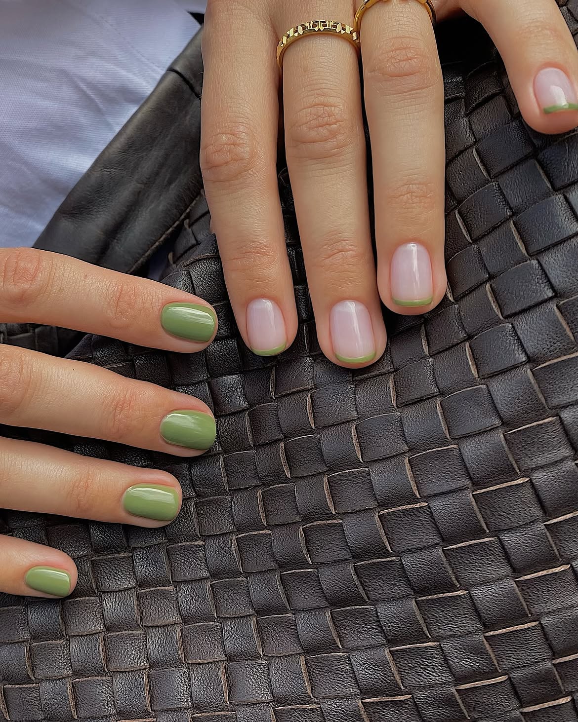 Duas m&atilde;os femininas sobre uma bolsa de couro tran&ccedil;ado marrom escuro. A m&atilde;o esquerda tem unhas curtas pintadas de verde-oliva. A m&atilde;o direita tem unhas com base transparente e pontas verdes, usando dois an&eacute;is dourados no dedo anelar