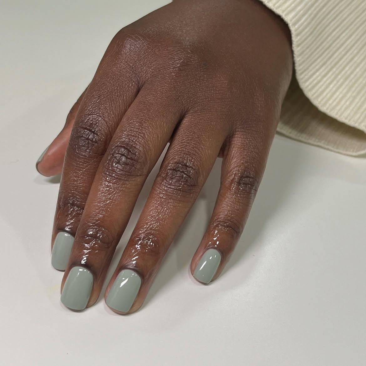 M&atilde;o de uma pessoa com pele escura, unhas curtas pintadas de verde-claro e cut&iacute;culas hidratadas, sobre uma superf&iacute;cie branca