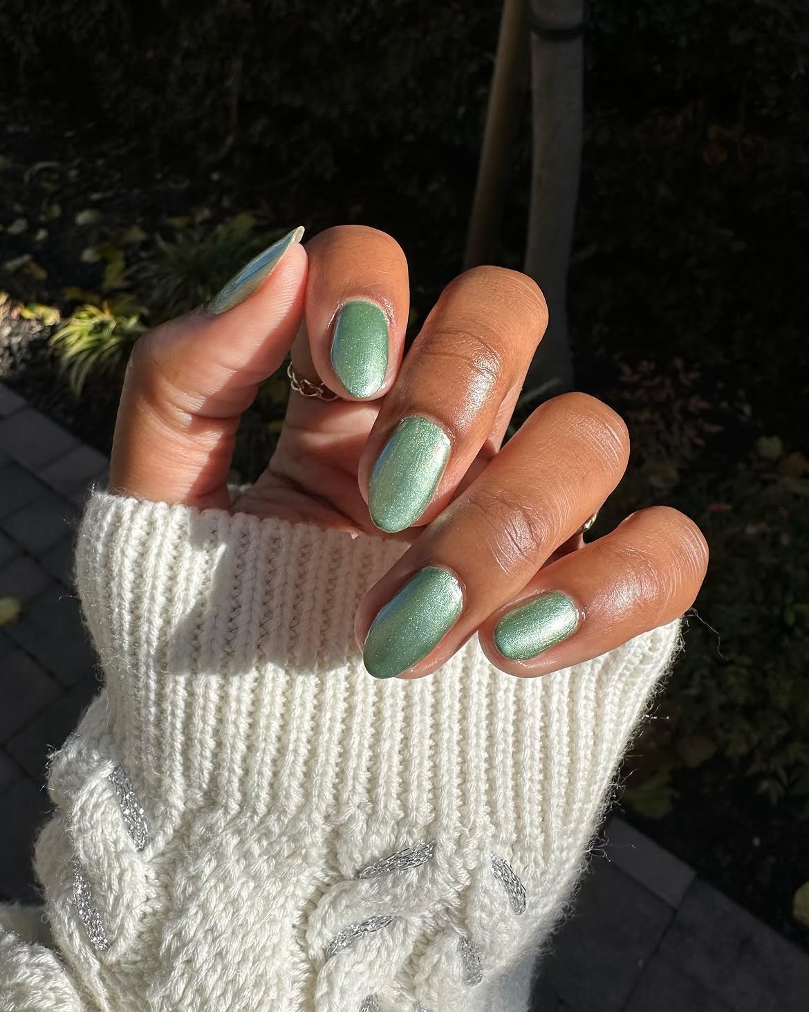 M&atilde;o de pele escura com unhas pintadas de verde met&aacute;lico, usando um anel dourado fino e uma blusa de tric&ocirc; creme com detalhes prateados, em ambiente externo com luz solar