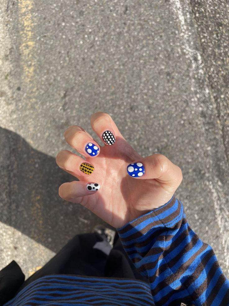 M&atilde;o feminina com unhas pintadas em bolinhas coloridas: azul com bolinhas brancas, preto com bolinhas brancas, amarelo com bolinhas pretas e branco com bolinhas pretas. A pessoa veste uma blusa de listras azuis e pretas, sobre um fundo de asfalto cinza com uma linha branca e sombra escura