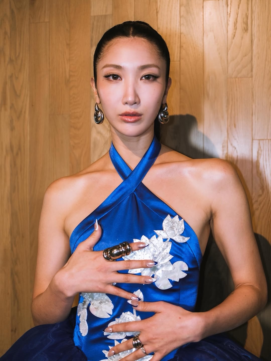 Mulher asi&aacute;tica com cabelo escuro preso, usando um vestido azul royal de al&ccedil;as cruzadas no pesco&ccedil;o e detalhes florais brancos. Ela tem brincos prateados grandes, an&eacute;is volumosos e unhas com pontas brancas e detalhes azuis. As m&atilde;os est&atilde;o sobre o peito e abd&ocirc;men, e o fundo &eacute; uma parede de madeira clara