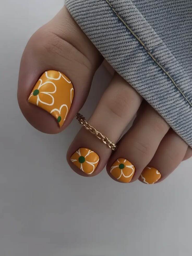 P&eacute; com unhas pintadas de laranja, decoradas com flores brancas e miolo verde, usando um anel de corrente dourado no segundo dedo. Uma cal&ccedil;a jeans azul clara aparece na parte superior da imagem