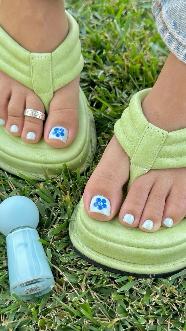 P&eacute;s com sand&aacute;lias verde-lim&atilde;o em um gramado. As unhas est&atilde;o pintadas de branco, com flores azuis nos ded&otilde;es. Um anel prateado adorna o segundo dedo do p&eacute; esquerdo. Um frasco de esmalte azul-claro est&aacute; no canto inferior esquerdo