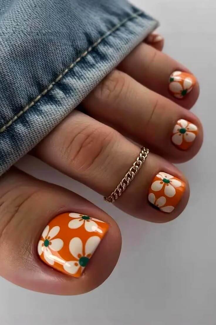 P&eacute;s com unhas pintadas de laranja vibrante e flores brancas com miolo verde, um anel dourado de corrente no segundo dedo, e a barra de uma cal&ccedil;a jeans azul clara na parte superior