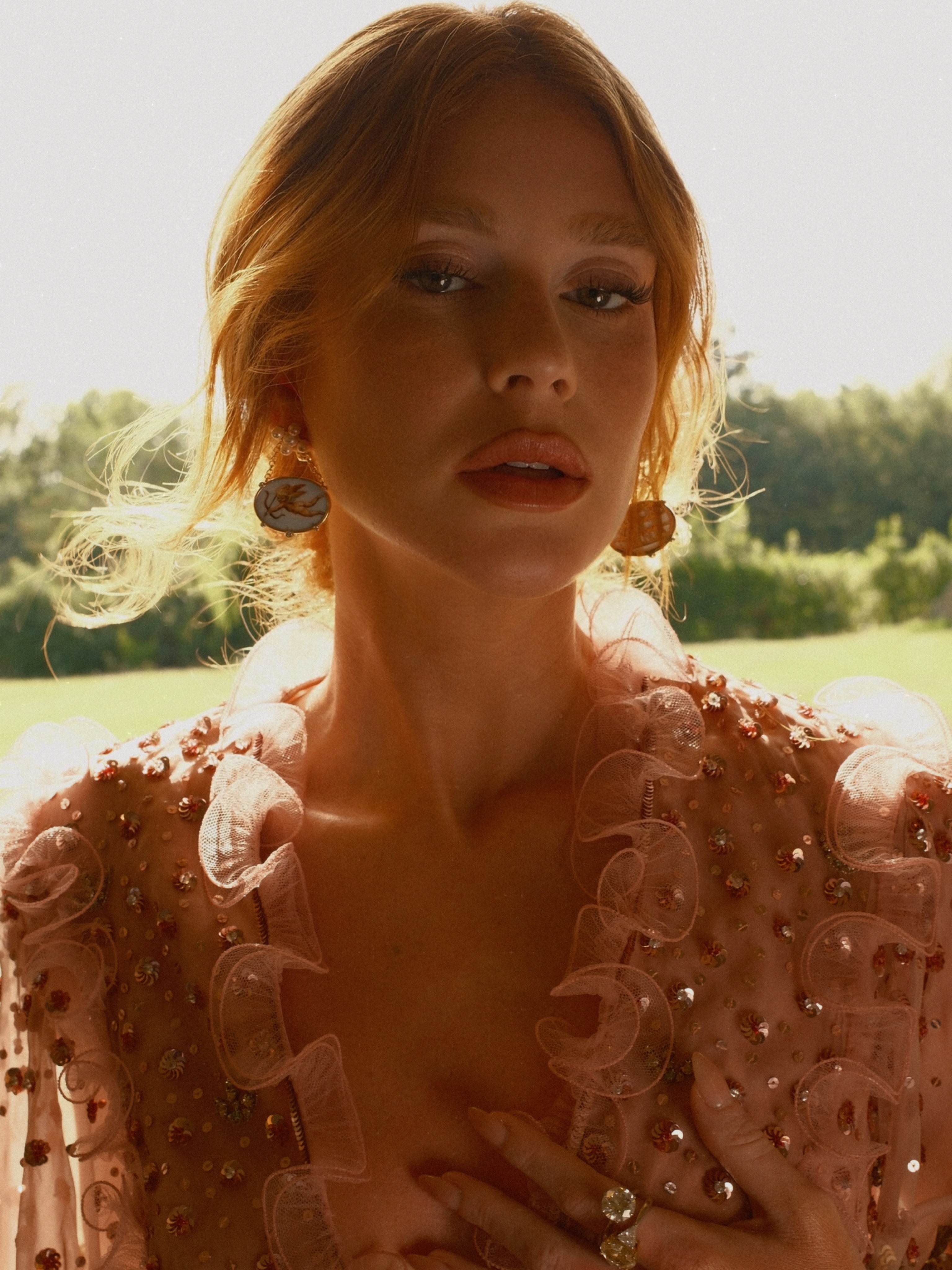 Mulher ruiva com maquiagem suave e l&aacute;bios rosados, usando brincos grandes e um vestido rosa transparente com babados e lantejoulas, em um cen&aacute;rio externo com vegeta&ccedil;&atilde;o verde e luz solar forte