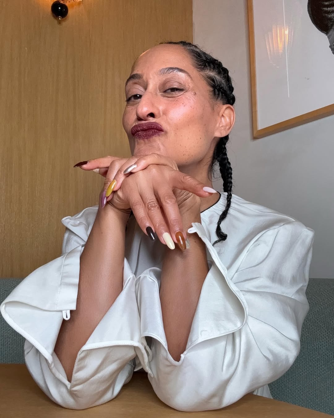 Tracee Ellis Ross, com tran&ccedil;as finas, faz bico e apoia o queixo nas m&atilde;os. Ela usa batom vinho, blusa branca de mangas bufantes e unhas longas coloridas em tons terrosos e met&aacute;licos. O fundo tem pain&eacute;is de madeira e um quadro