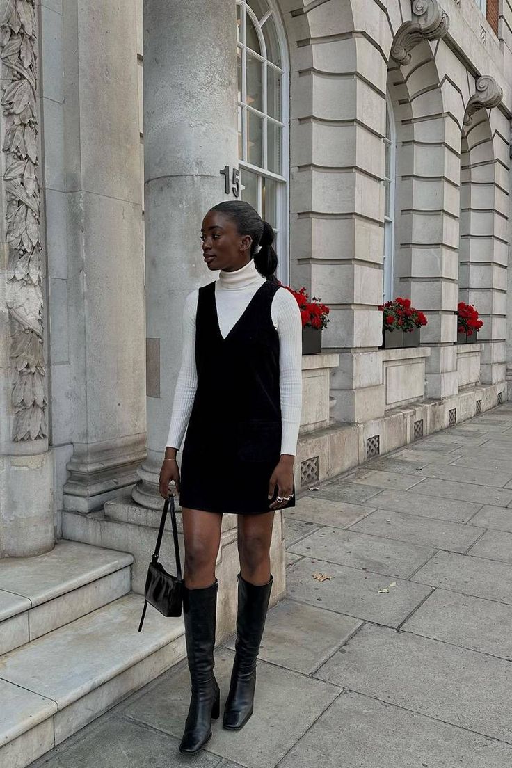 Mulher negra com cabelo preso, usando um vestido preto sobre uma blusa de gola alta branca e botas pretas de cano alto, segurando uma bolsa preta. Ela est&aacute; em uma cal&ccedil;ada de pedra, ao lado de um pr&eacute;dio cinza com janelas arqueadas e vasos de flores vermelhas. O n&uacute;mero 15 est&aacute; vis&iacute;vel na parede do pr&eacute;dio
