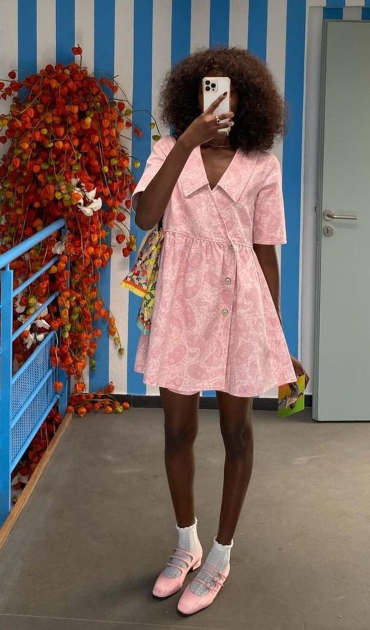 Mulher de pele escura e cabelo crespo, usando um vestido rosa claro com estampa paisley e gola pontuda, tira uma selfie com um celular branco. Ela veste meias brancas e sapatos Mary Jane rosa com fivelas. Ao fundo, uma parede com listras azuis e brancas, uma porta cinza e um arranjo de flores alaranjadas em um corrim&atilde;o azul