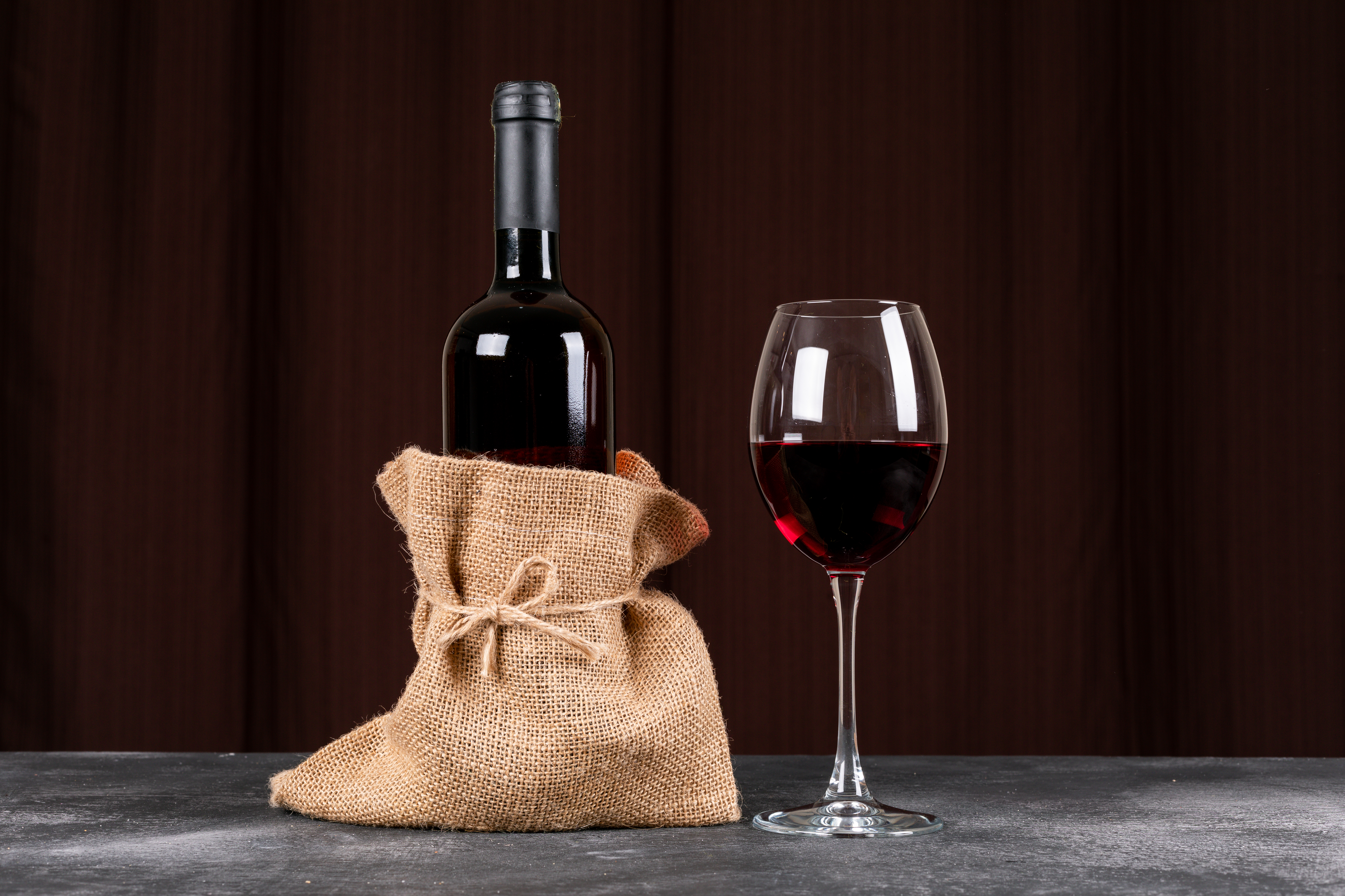 Garrafa de vinho tinto em saco de juta amarrado, ao lado de uma taça de vinho cheia, sobre superfície escura com fundo marrom