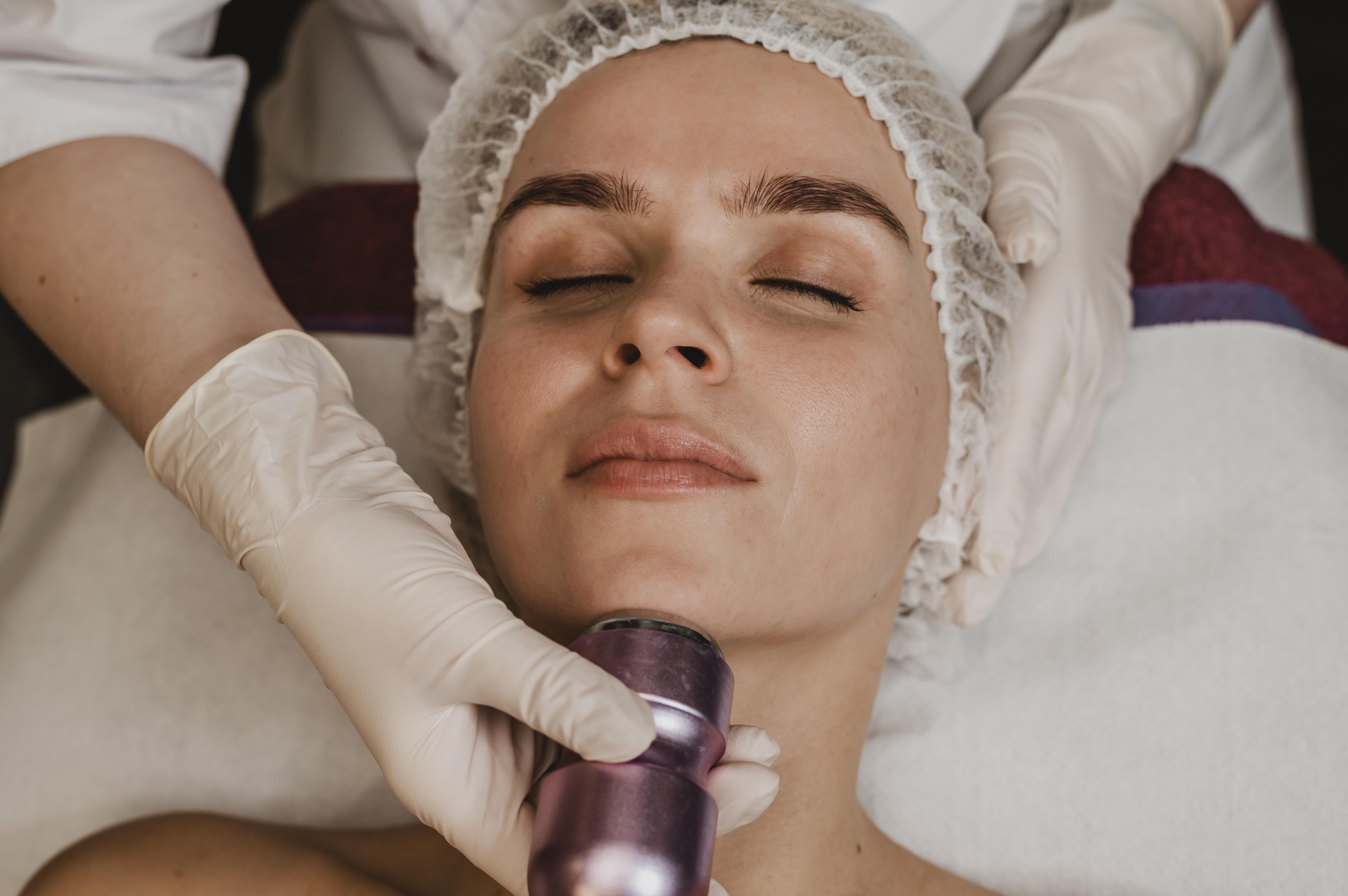 Mulher deitada com touca descart&aacute;vel e olhos fechados, recebendo tratamento est&eacute;tico no queixo com aparelho roxo, aplicado por m&atilde;os com luvas brancas