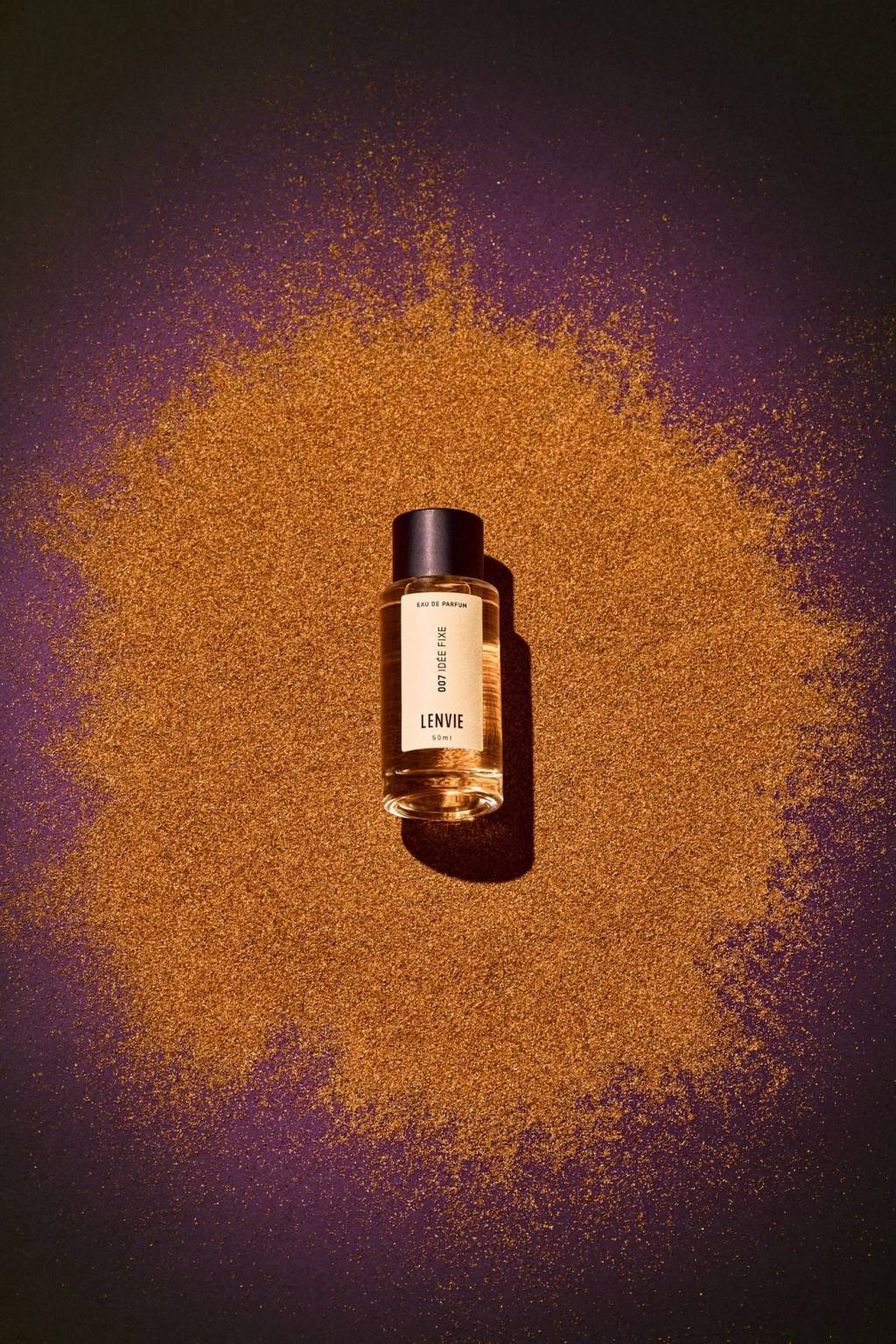 Frasco de perfume Lenvie 007 Id&eacute;e Fixe de 50ml, com l&iacute;quido &acirc;mbar e tampa preta, sobre uma superf&iacute;cie roxa escura coberta por glitter dourado em formato de nuvem