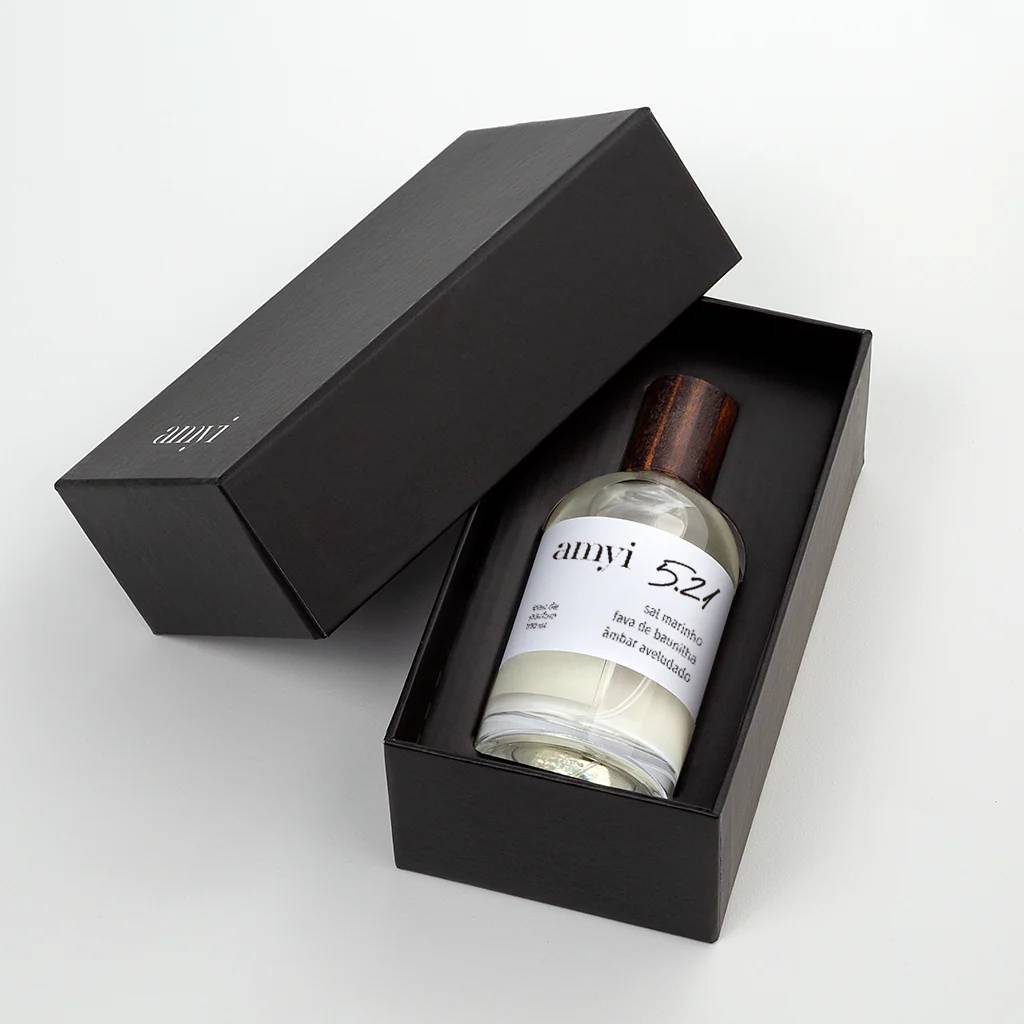 Frasco de perfume Amyi 5.21 com tampa de madeira escura dentro de uma caixa preta aberta, sobre fundo branco