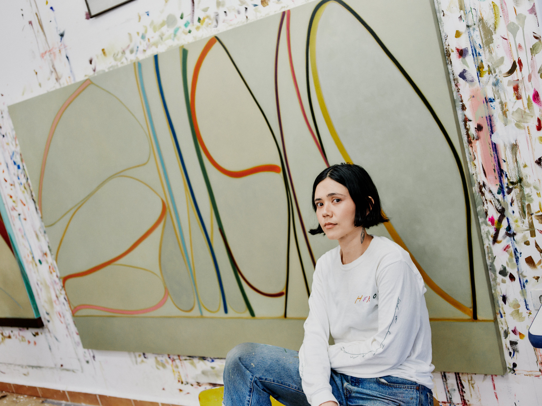 Mulher de cabelo preto curto, camiseta branca e jeans, sentada em um ateliê de arte, com uma grande tela abstrata de fundo e paredes respingadas de tinta