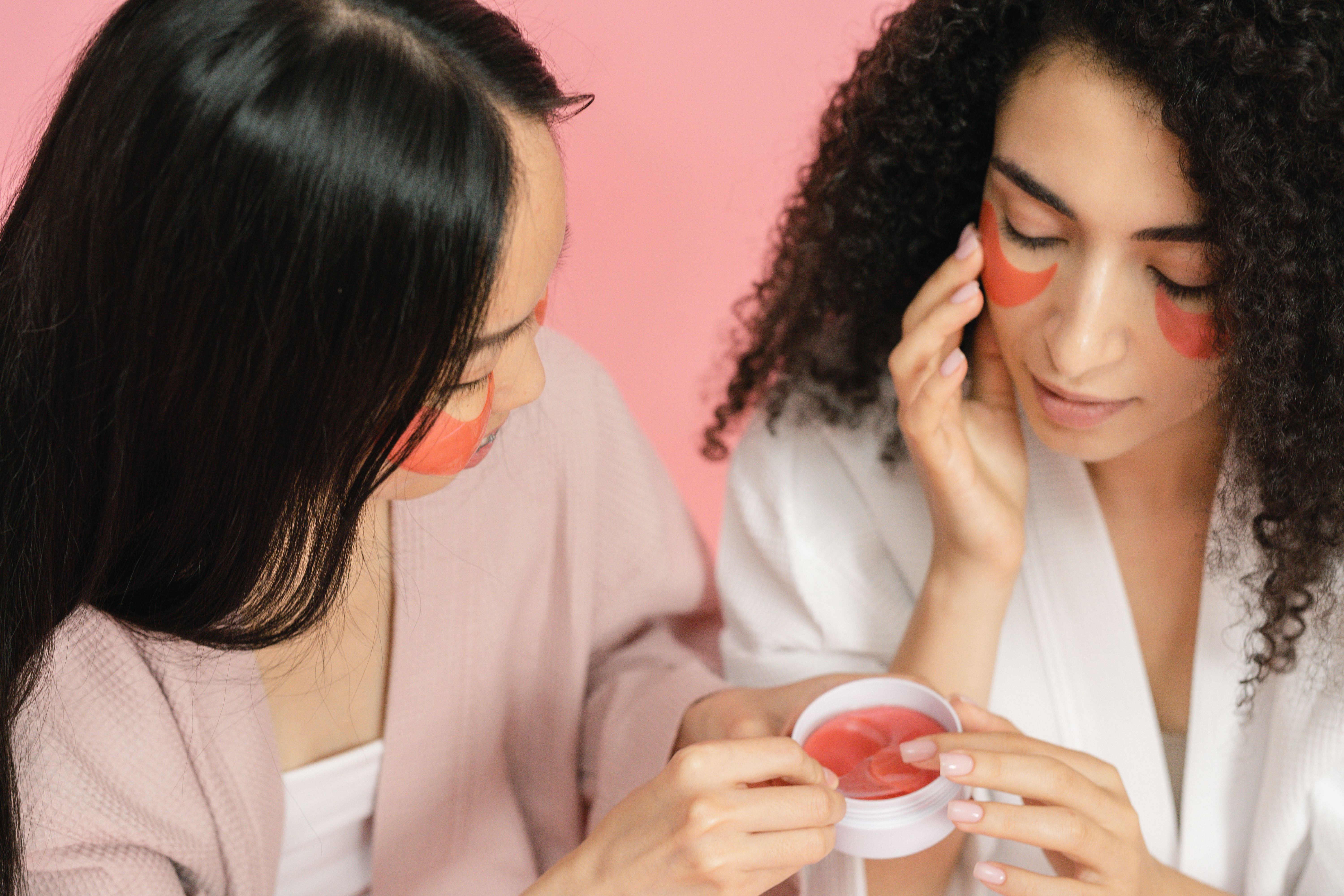 Duas mulheres com máscaras faciais de hidrogel sob os olhos. Uma mulher de cabelos lisos e escuros, usando roupão rosa, aplica uma máscara. A outra, de cabelos cacheados e escuros, com roupão branco, já tem as máscaras aplicadas e toca o rosto, com os olhos fechados, em fundo rosa